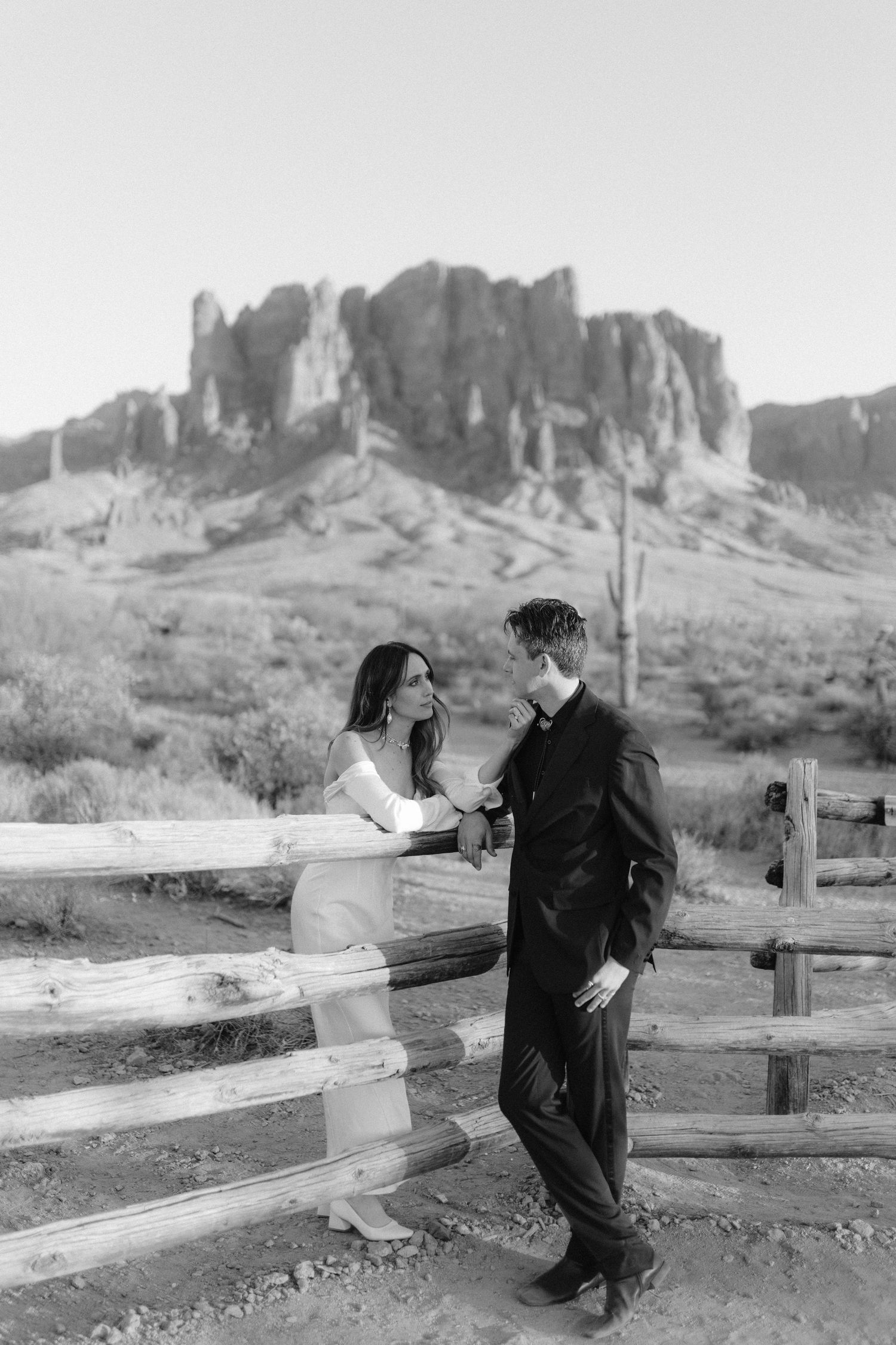 Black and white photo of couple sharing a moment by a wooden fence with Superstition Mountains backdrop.