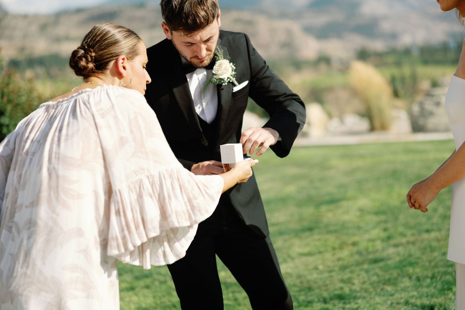 Wedding coordinator adjusting groom's cufflinks in tuxedo outdoors with mountain backdrop.