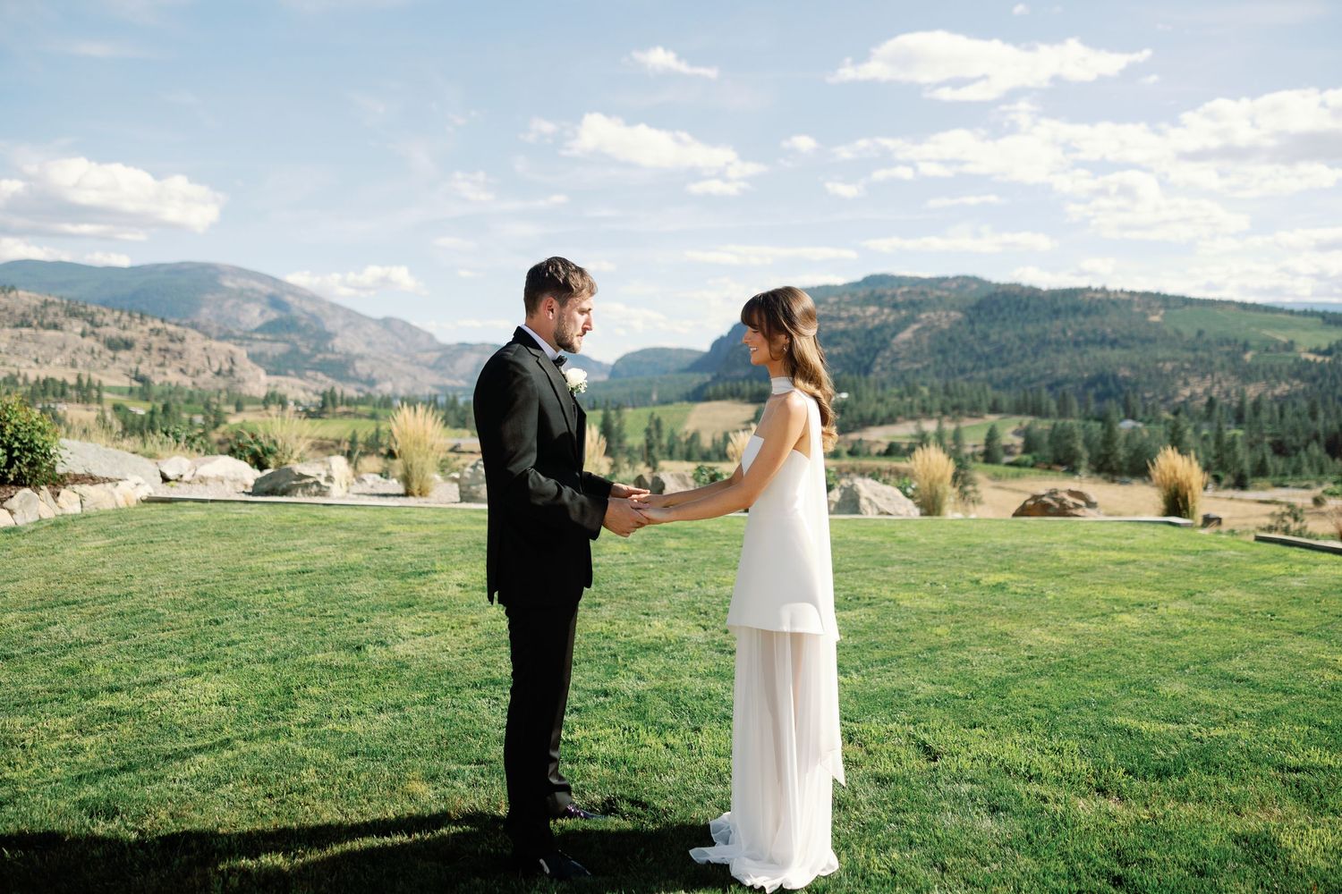 Bride and groom hold hands facing each other on a lush lawn with scenic mountain backdrop.