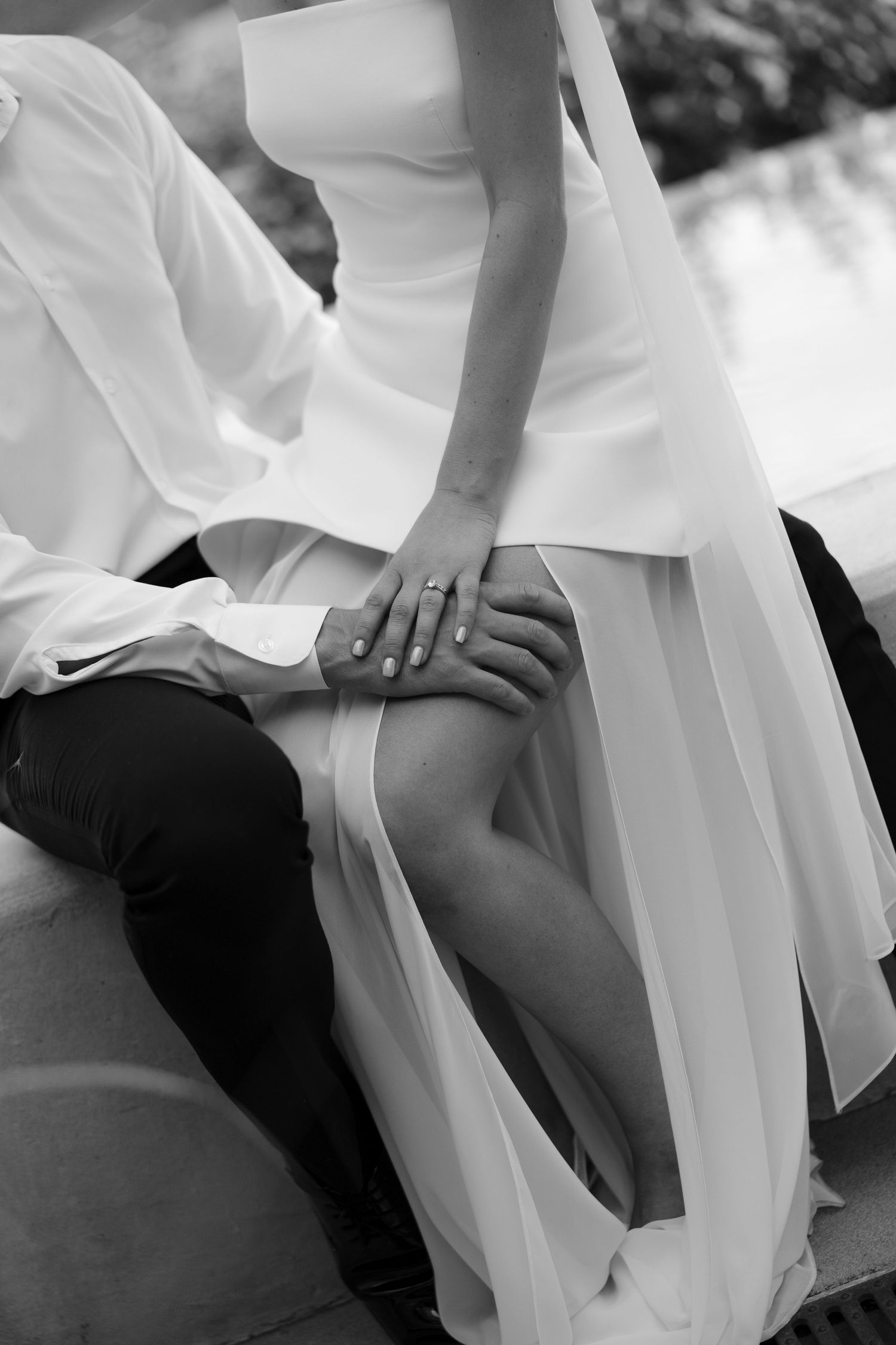 Close-up black and white photo of bride and groom holding hands, showcasing wedding ring and elegant bridal gown.