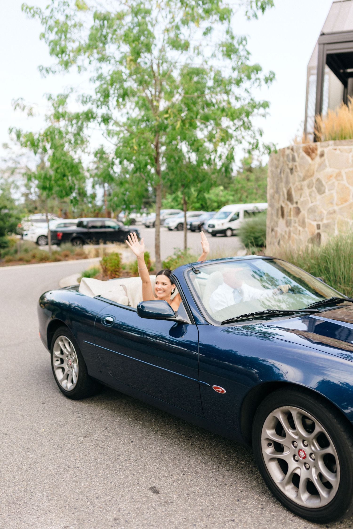 Woman with raised arms smiling in a blue convertible sports car parked near a stone building with trees.