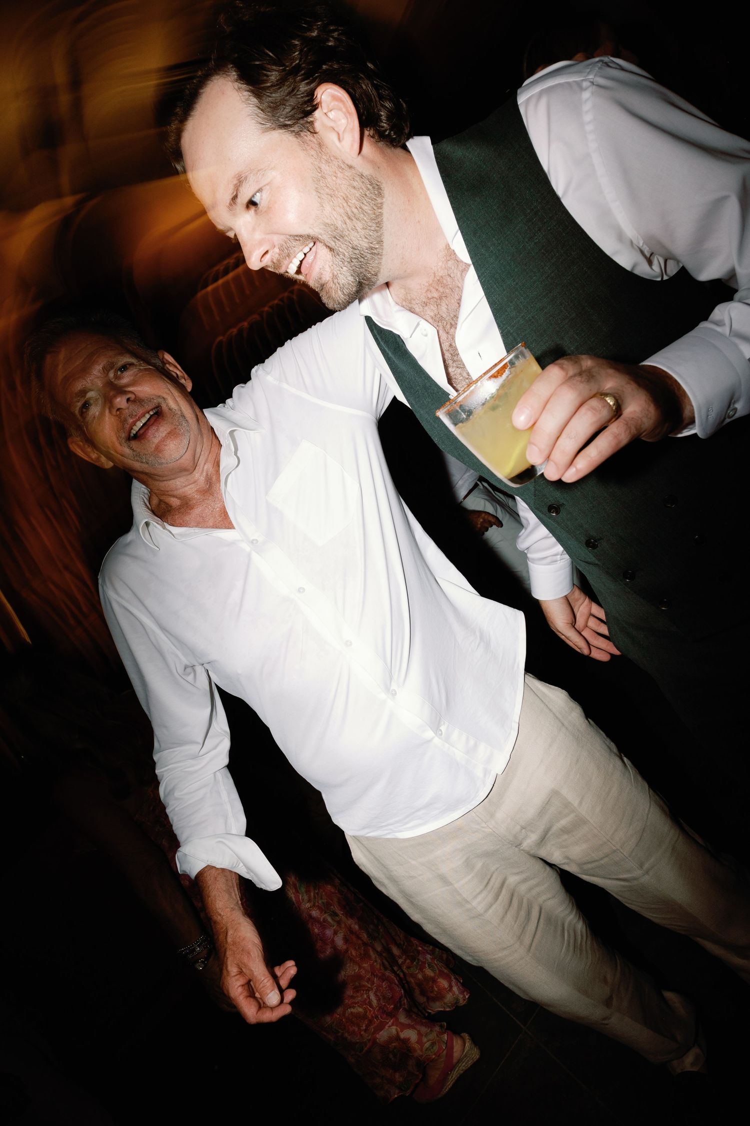 Two men laugh together at wedding reception, one holding a gold card.