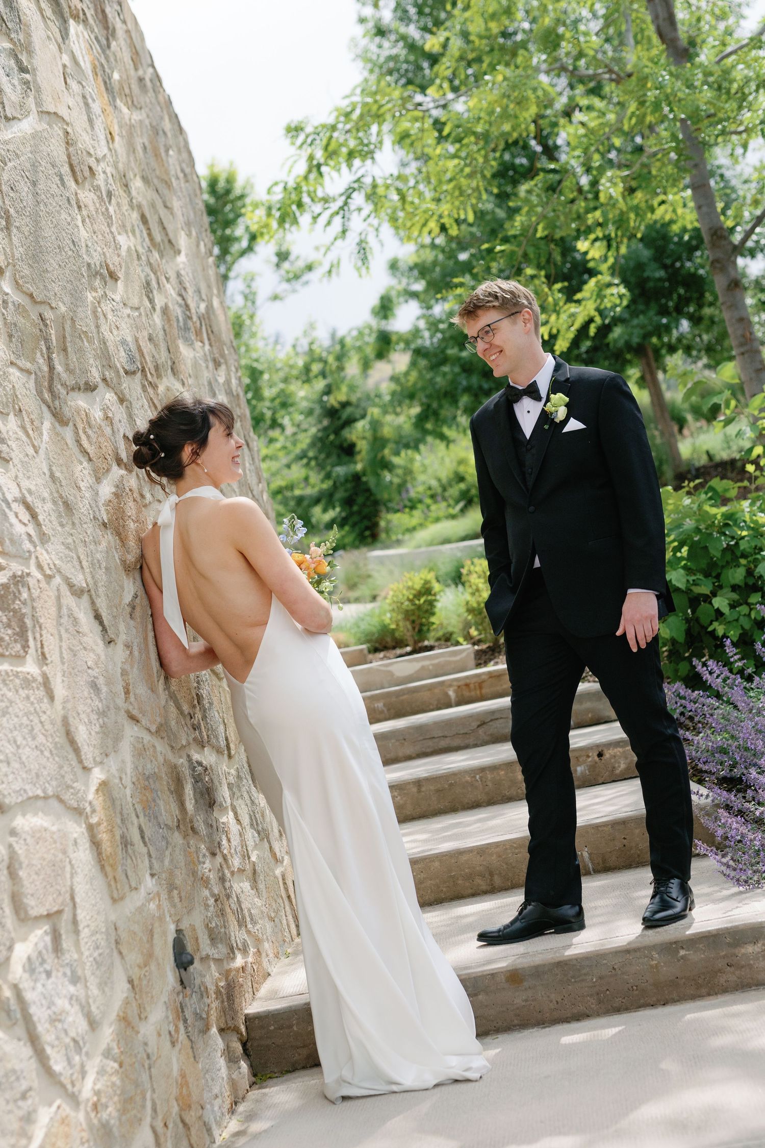Bride in backless white gown leans against stone wall as groom in black tuxedo smiles at her outdoors.