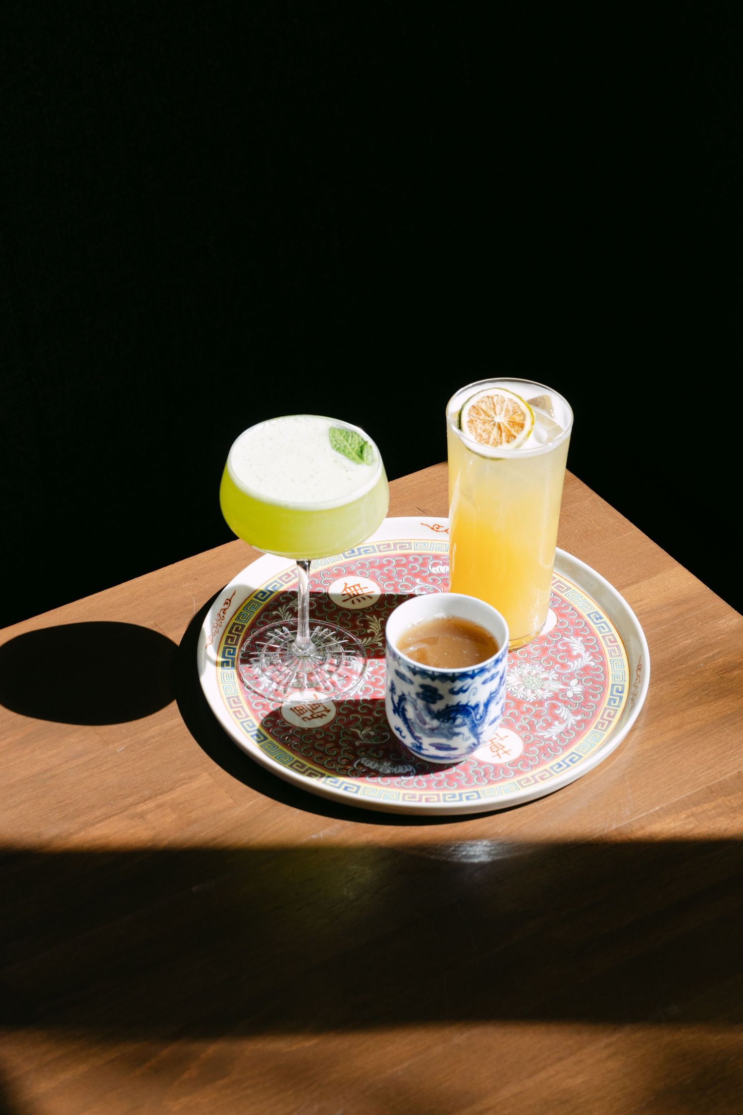 Three elegant cocktails on a decorative tray: a green foam drink, orange lemon highball, and small espresso cup on wooden table.