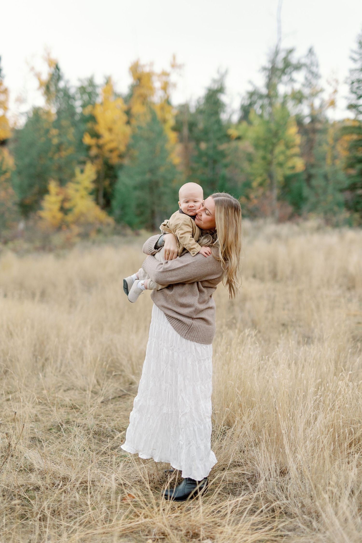 A mother in a white skirt and tan sweater lovingly kisses her baby while standing in a golden autumn meadow.