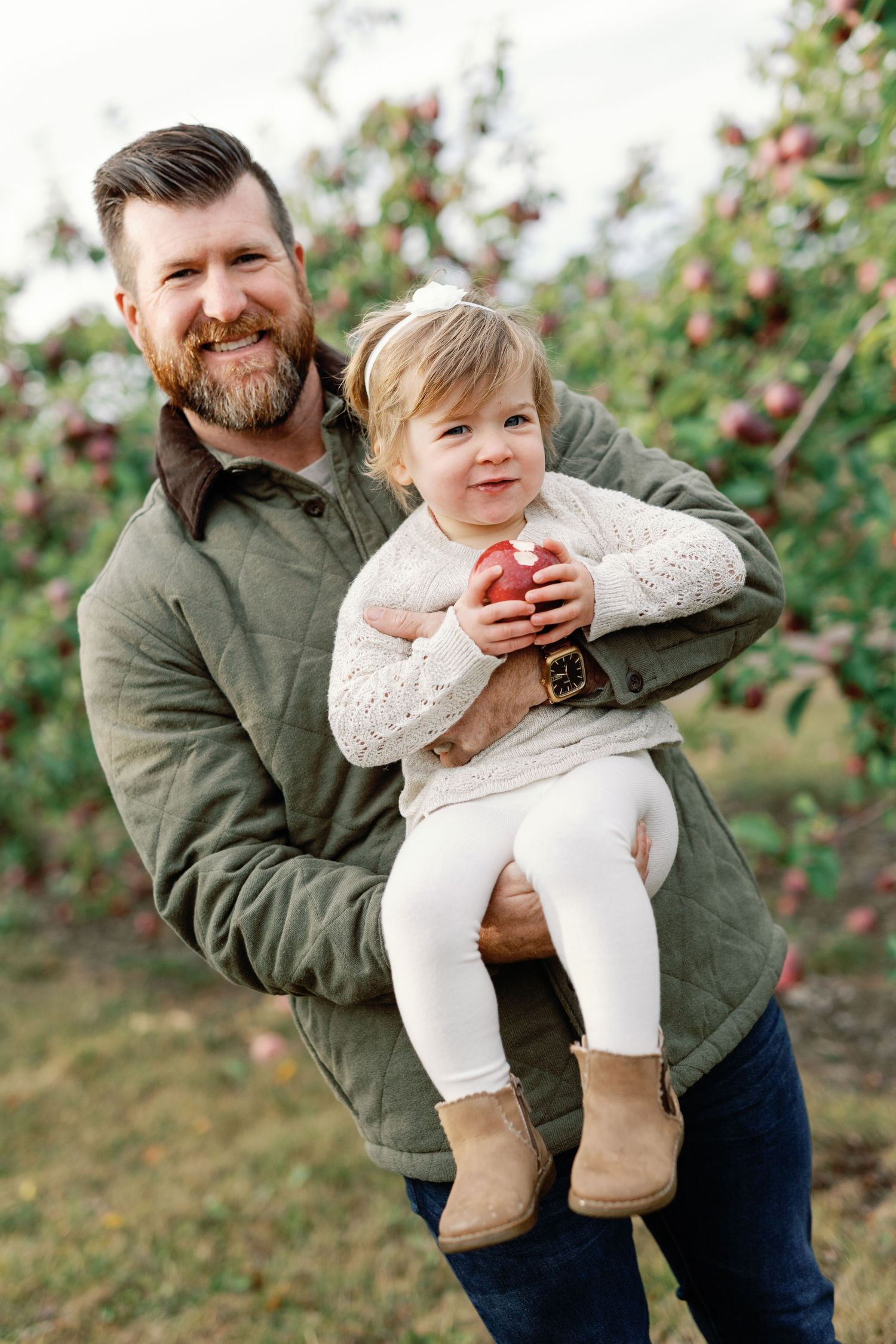 Father holding young daughter in apple orchard, child clutching red apple, both smiling outdoors in fall.