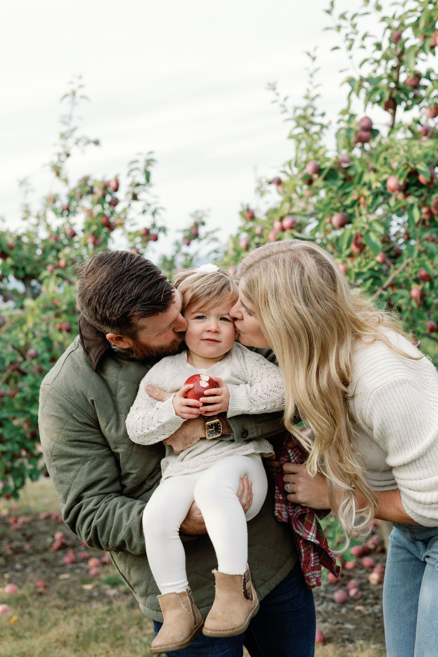 A family of three shares kisses with their toddler holding a red apple in an autumn orchard.