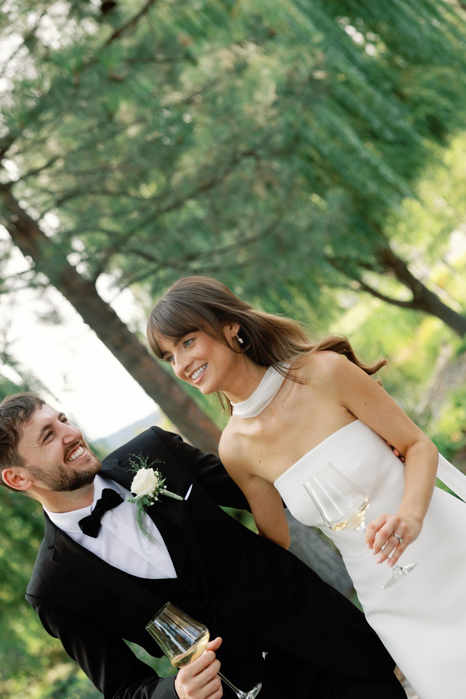 Smiling bride in white halter gown and groom in black tuxedo share champagne glasses under a pine tree.