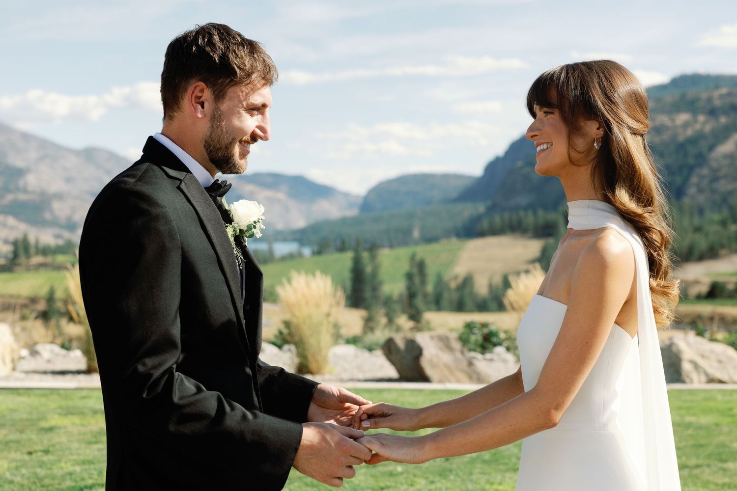 Smiling bride and groom exchange rings during sunny outdoor wedding with mountain scenery.
