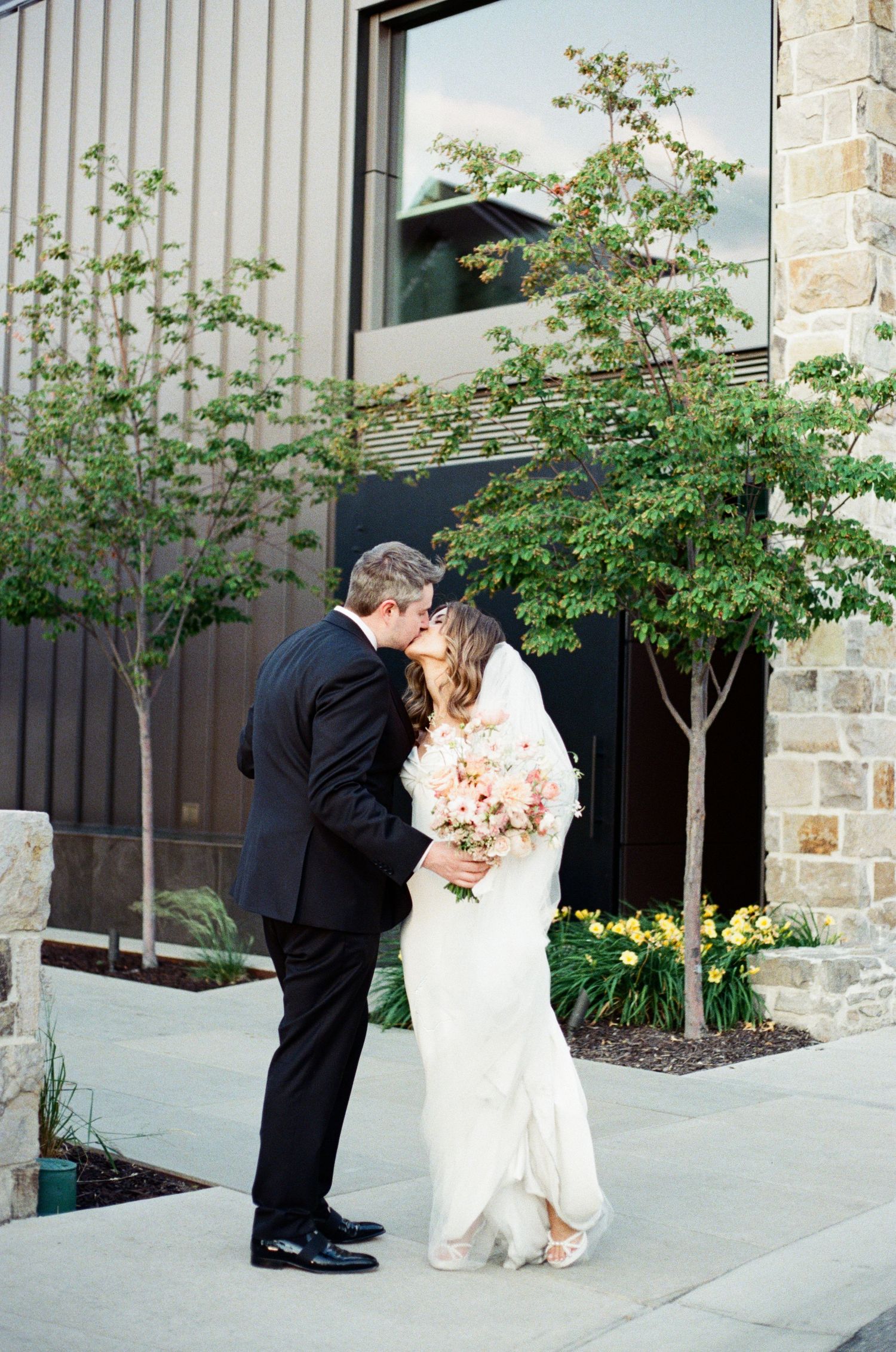 Bride and groom share a romantic kiss outdoors, she holding a peach floral bouquet in a white gown.