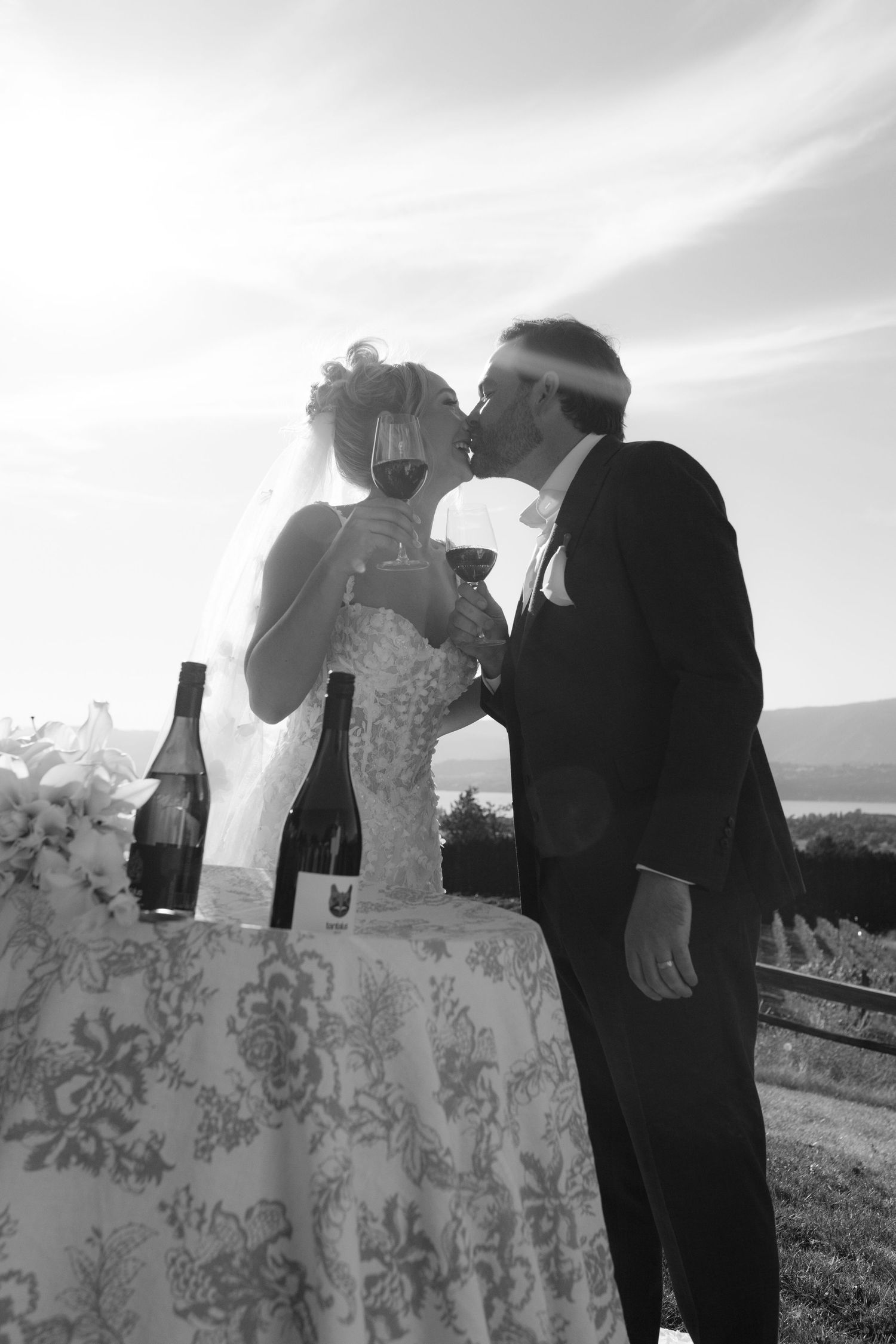 Black and white silhouette of newlyweds kissing while toasting with red wine at vineyard wedding.