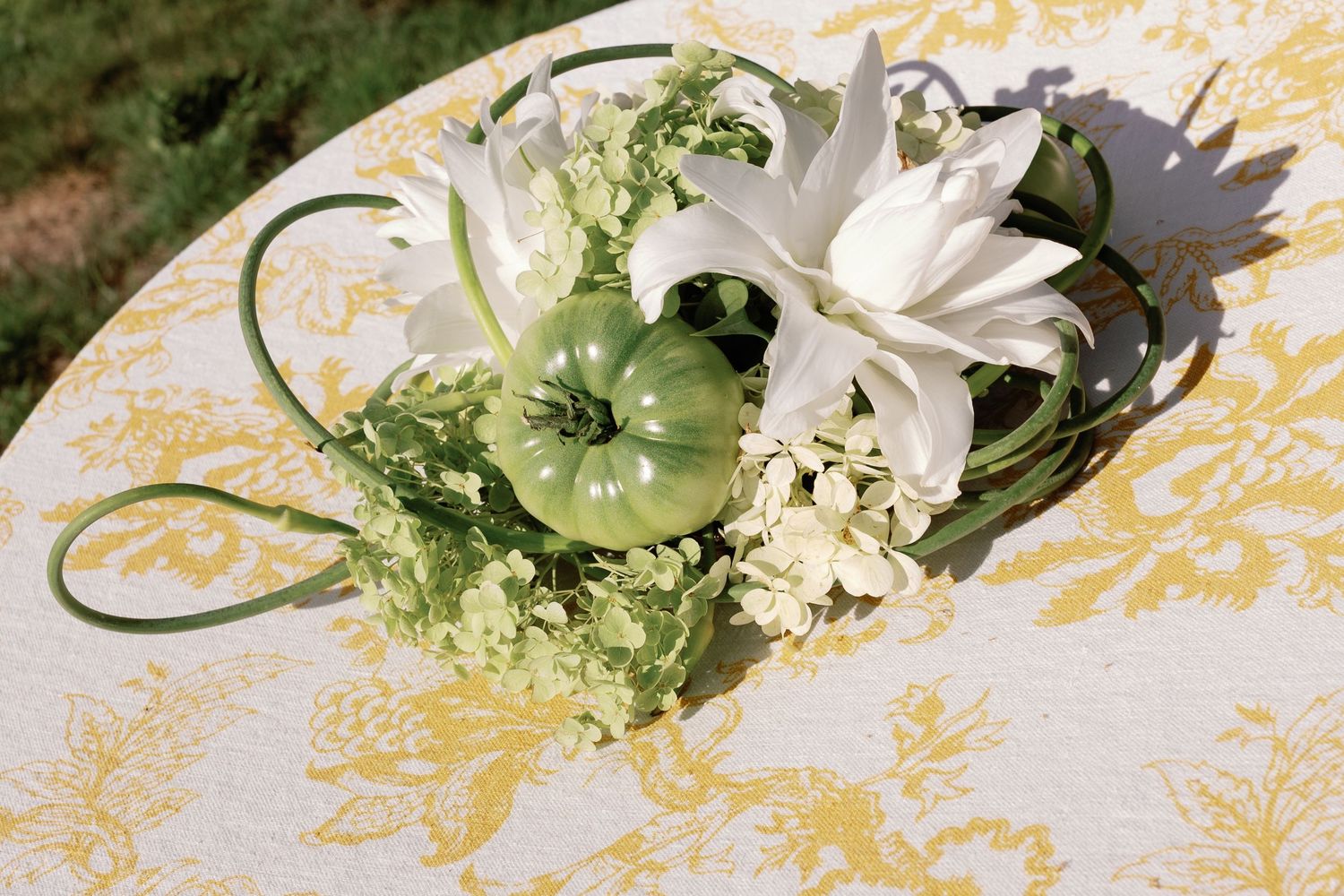 Elegant wedding centerpiece featuring white lilies, green tomatoes, and delicate blooms on yellow damask tablecloth.
