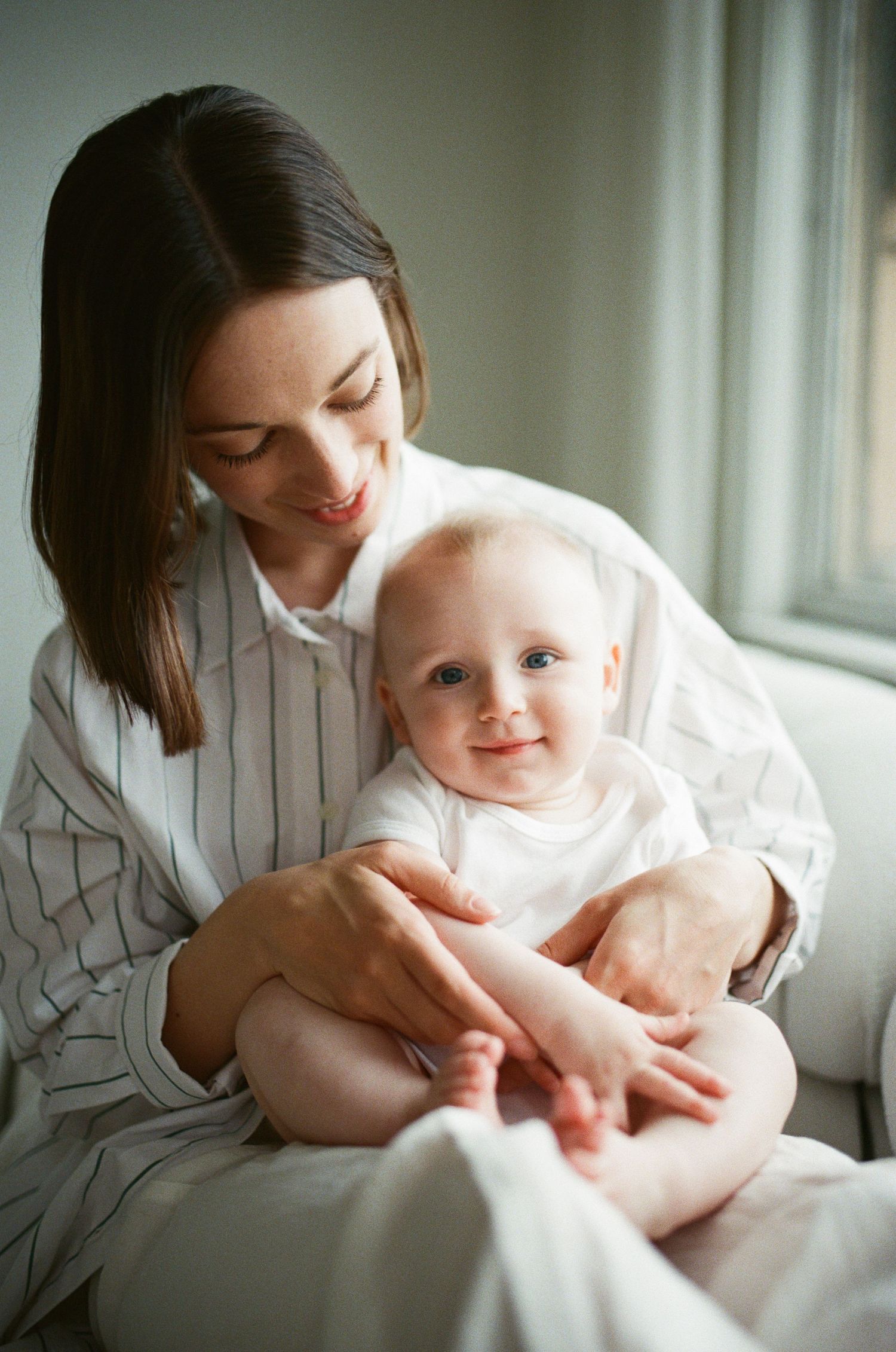 Smiling mother holding happy blue-eyed baby near bright window indoors.
