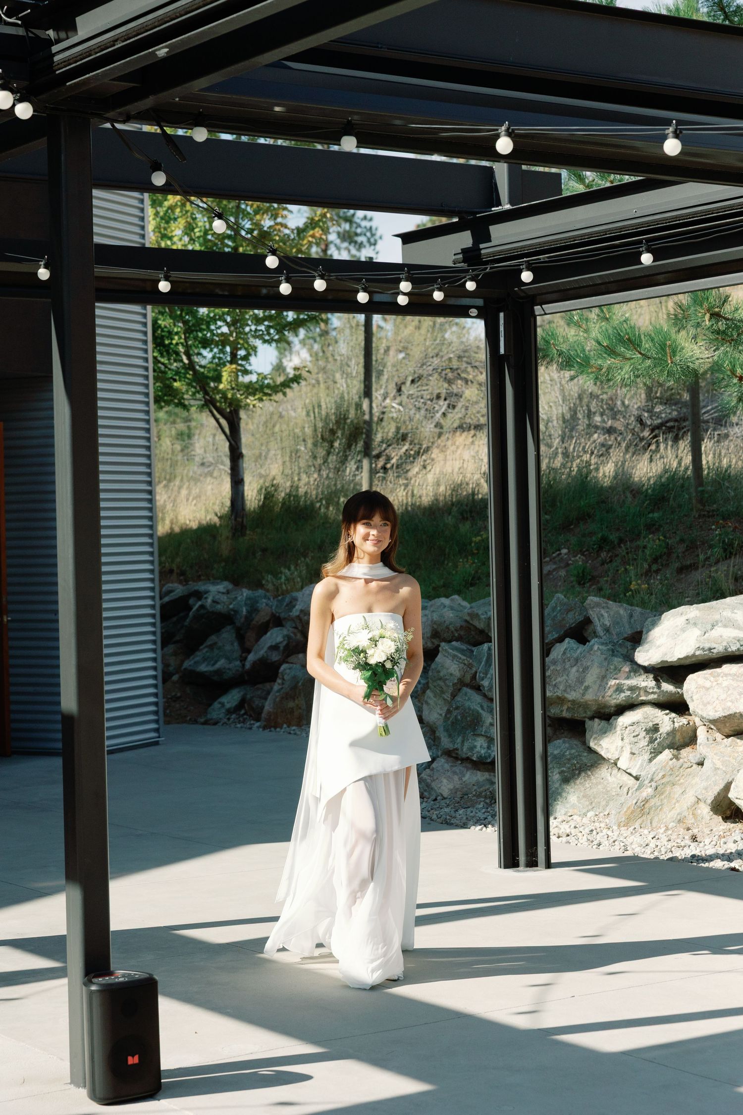 Elegant bride in strapless white gown holds bouquet under modern pergola with string lights.