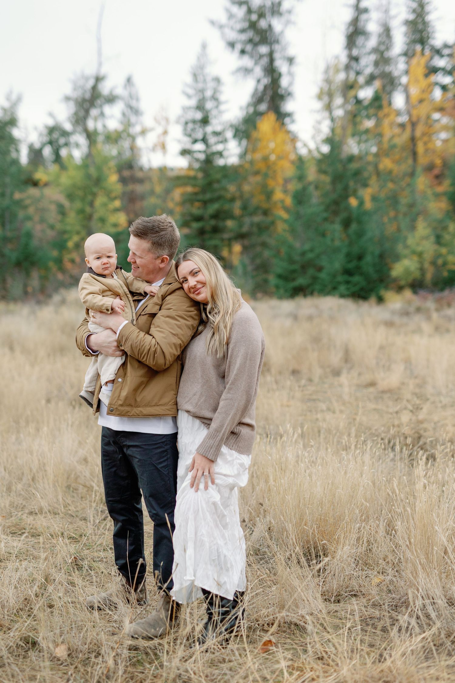 A family poses in an autumn field, father holding baby while mother leans close, surrounded by golden grass and colorful trees.