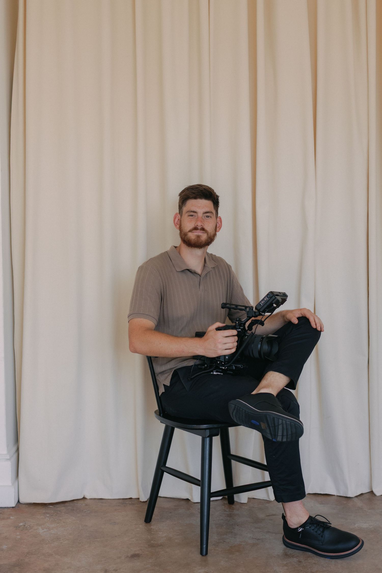 Man with beard sits on black chair holding professional camera, wearing tan polo and black pants against cream curtains.
