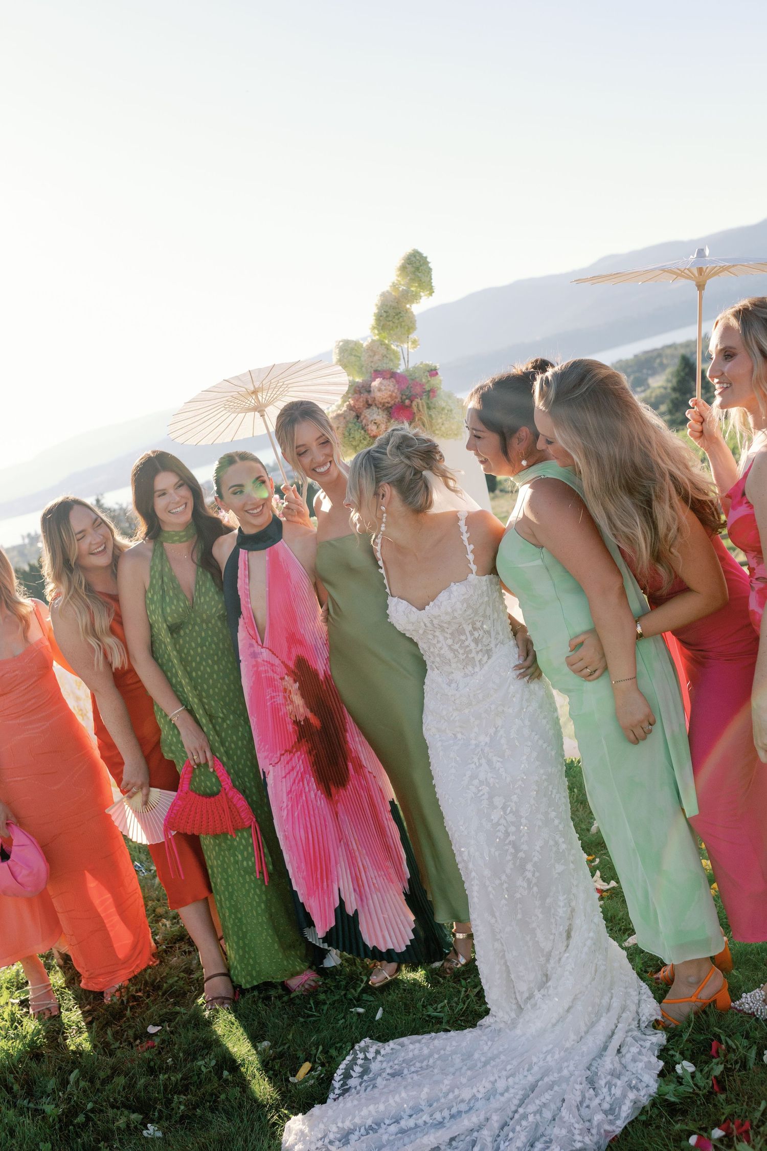 Bride in lace gown surrounded by bridesmaids in colorful green and pink dresses at outdoor wedding.