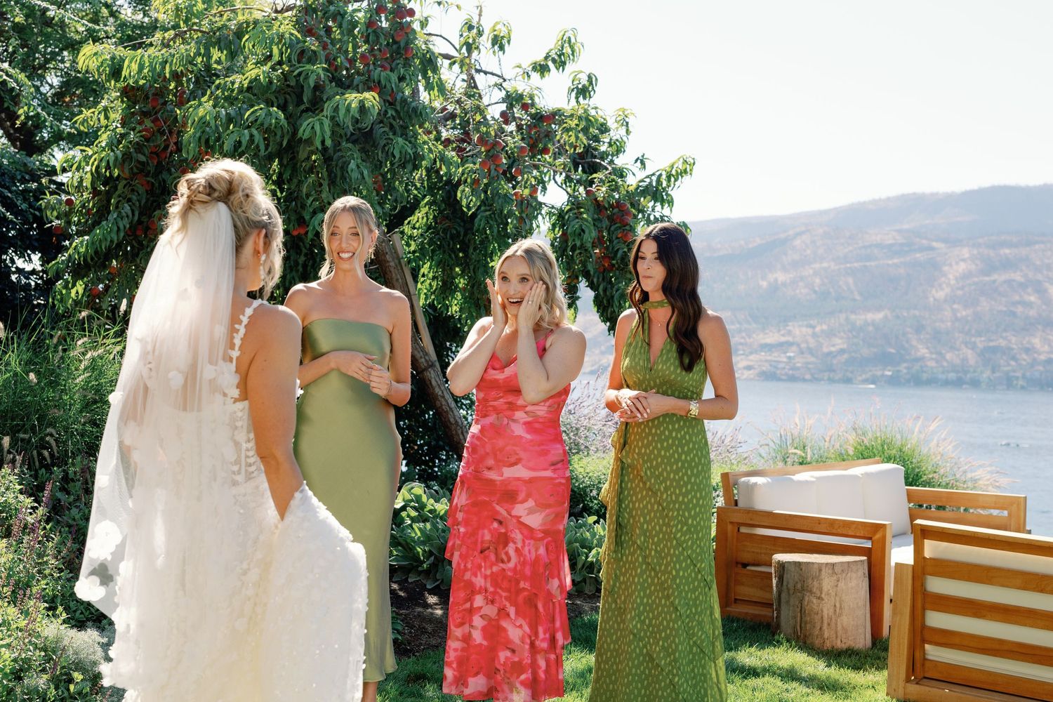 Bride in veil surprises three bridesmaids in colorful dresses with emotional first look in garden by lake.