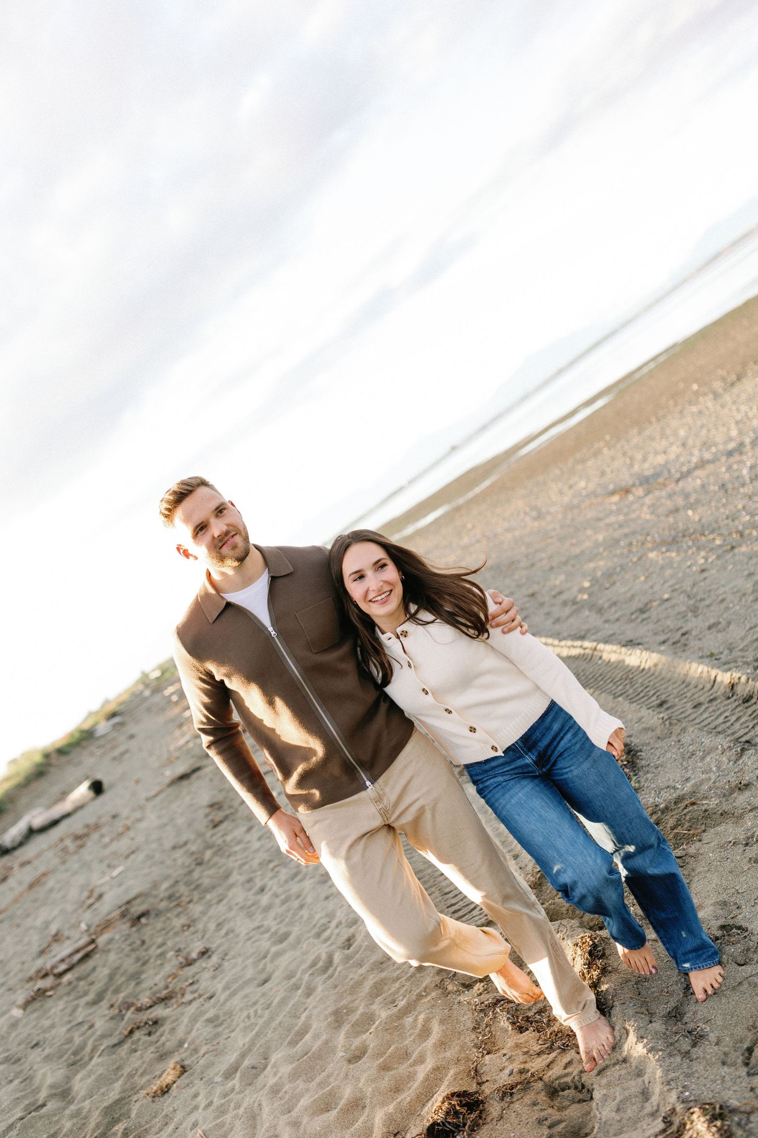 A happy couple walks barefoot on a sandy beach, the man in a brown jacket and the woman in a white sweater and jeans.
