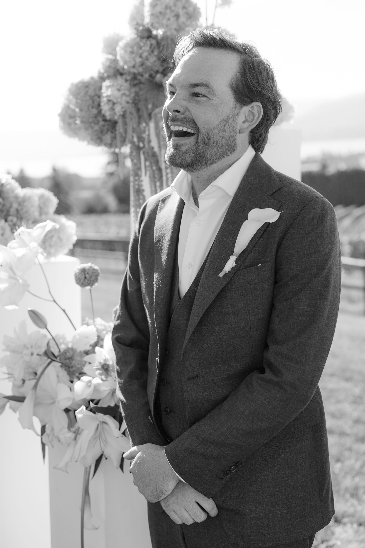 Black and white photo of laughing groom in suit at outdoor wedding ceremony altar.