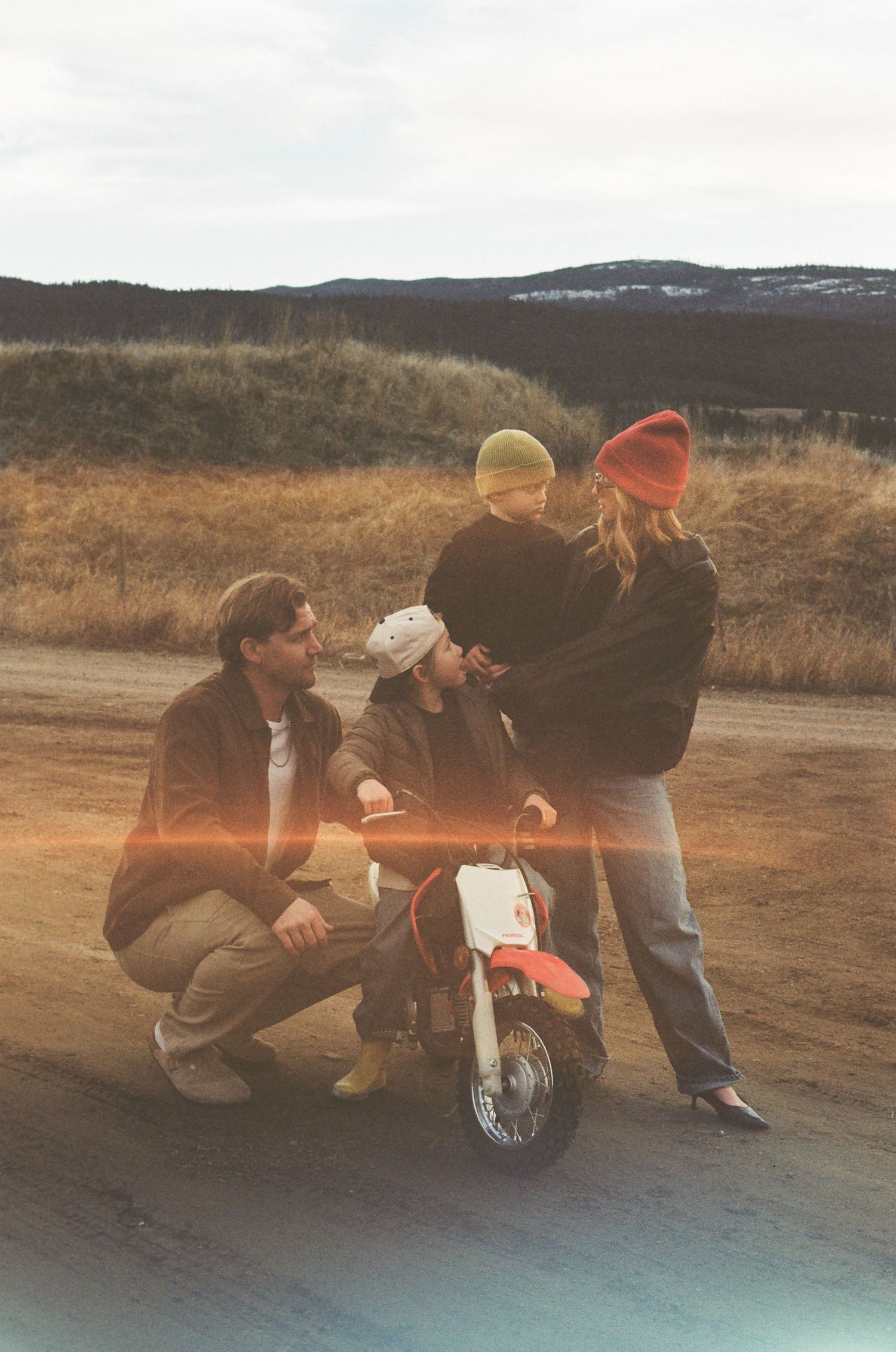 Family with two children gathered around a small motorcycle on a rural dirt road with mountains in the background.