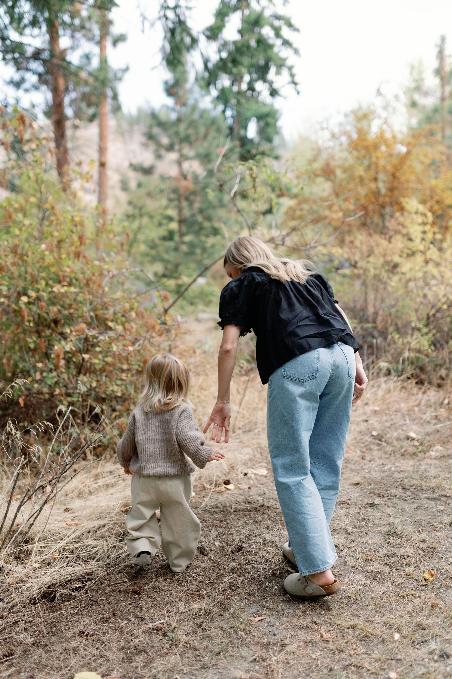 A mother reaches out to her toddler on a forest path surrounded by autumn trees and foliage.