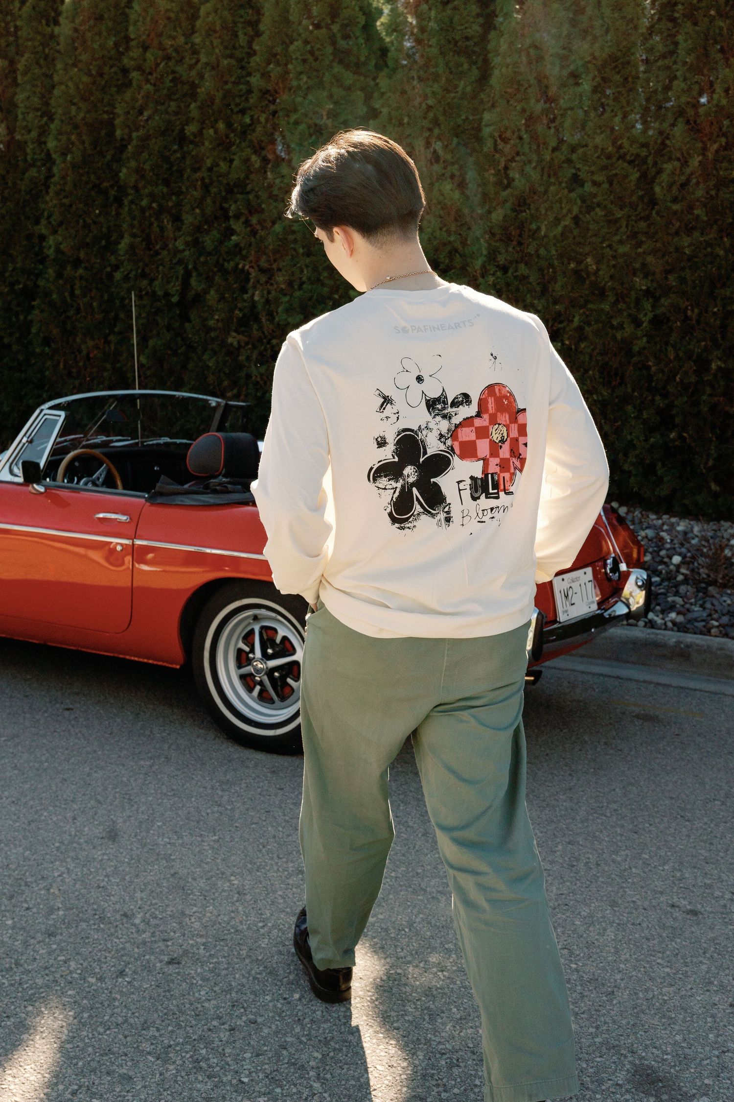 Young man in cream graphic long-sleeve shirt and sage green pants standing beside a classic red convertible car.