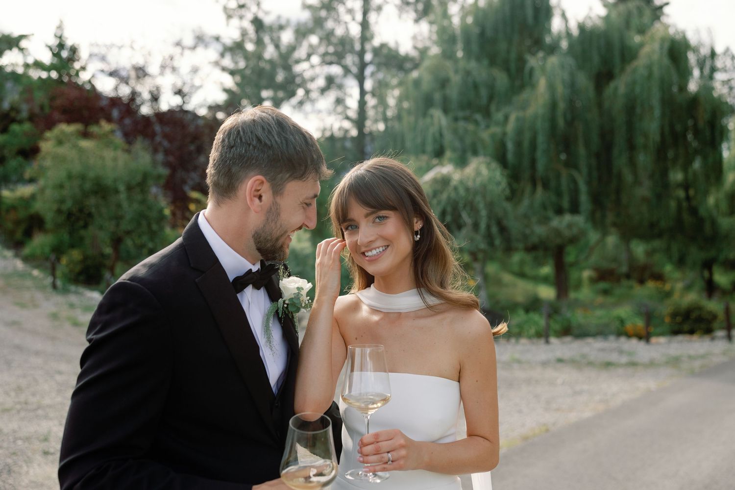 Bride in white halter gown smiles while groom in black tuxedo leans close on a tree-lined gravel path.