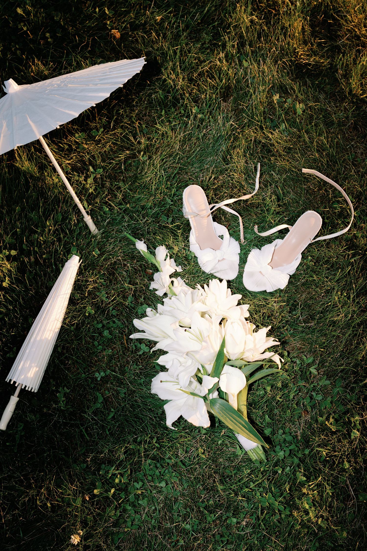 White tulip bridal bouquet with strappy heels and parasol laid on green grass for a wedding flat lay.