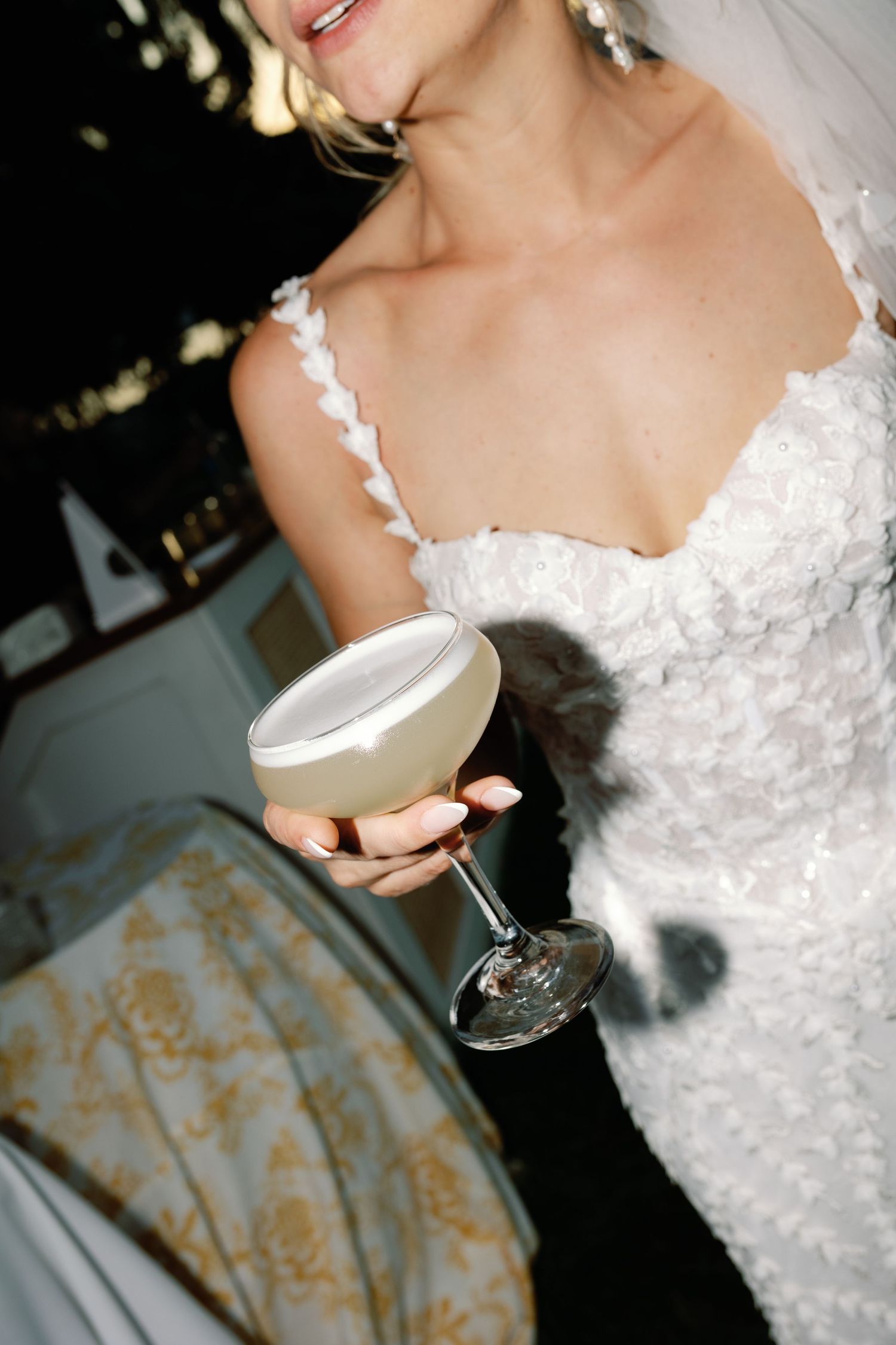 Bride in white floral wedding gown holding elegant coupe glass with frothy cocktail at reception.