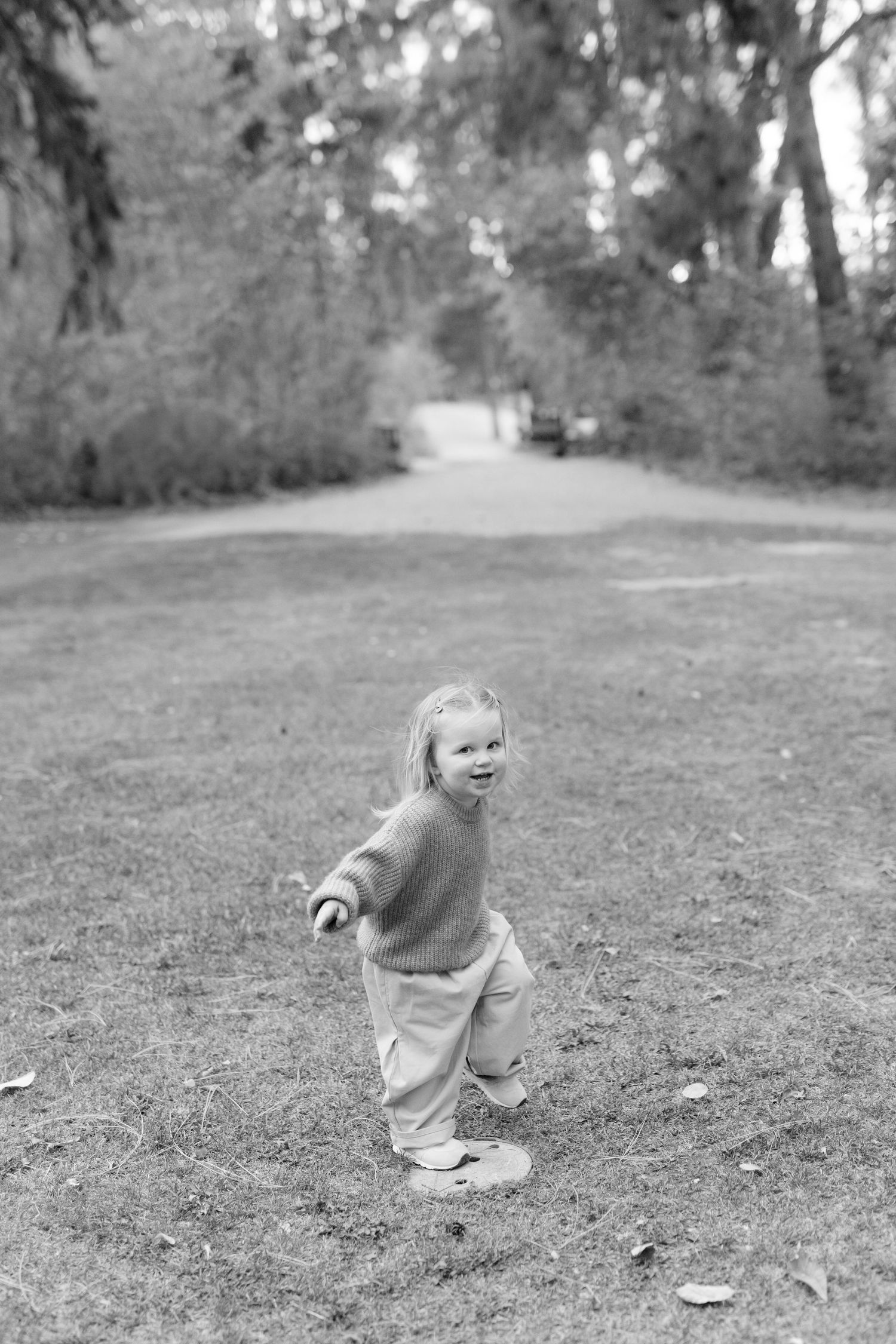 Black and white photo of a happy toddler running on grass in a park, wearing a sweater and pants.
