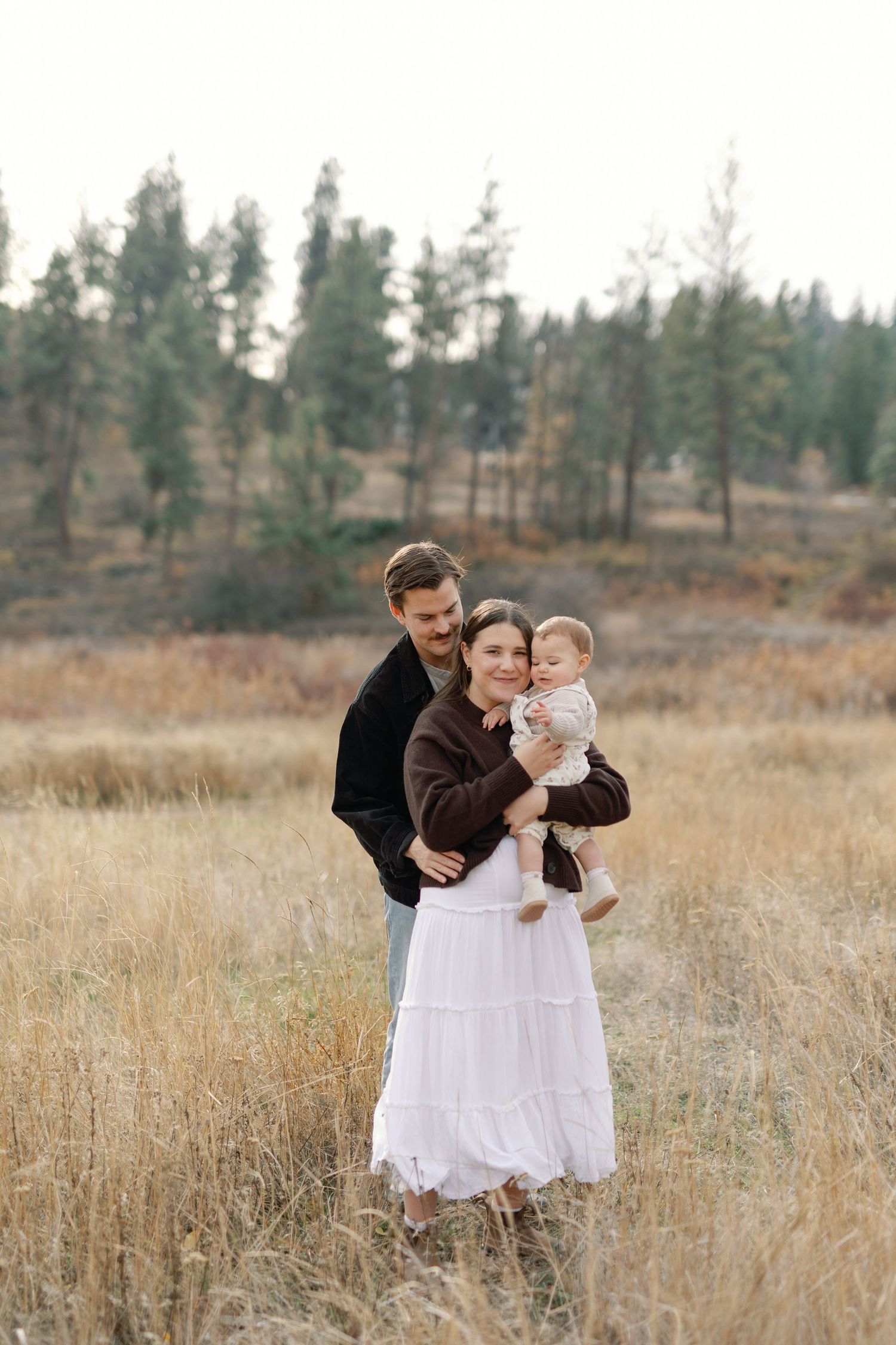 A young family poses in a golden field, parents holding their baby, with pine trees in the background.