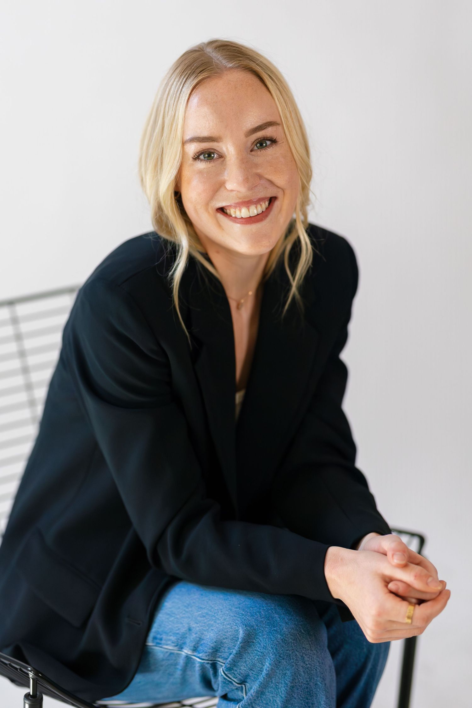 Smiling blonde woman in black blazer and jeans sitting relaxed with hands clasped in bright studio setting.