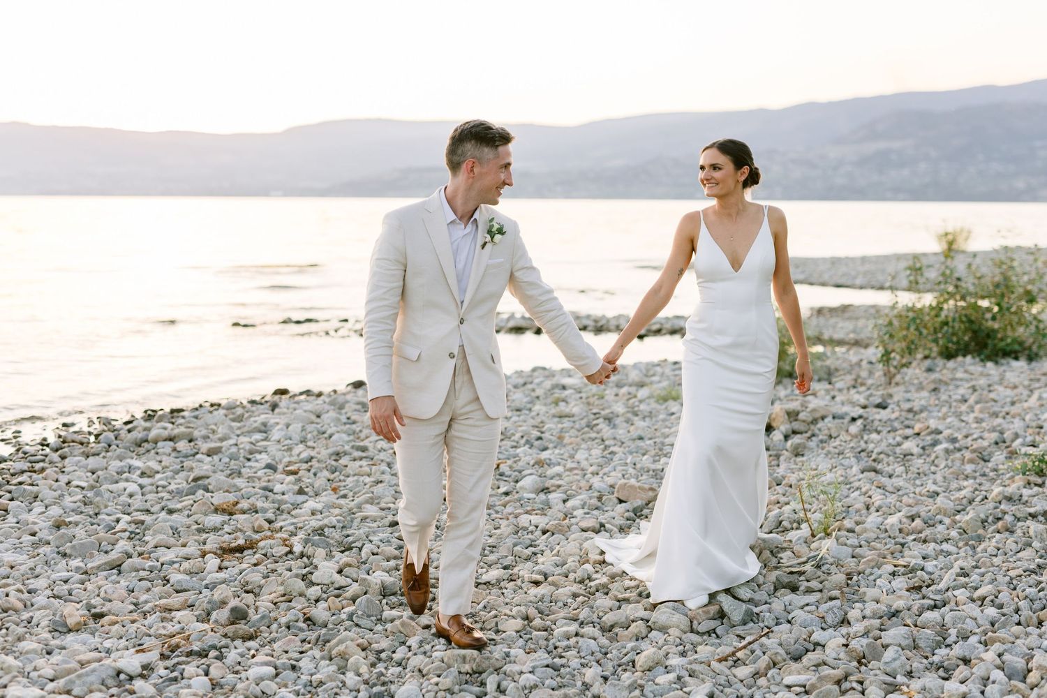 Bride and groom hold hands on rocky beach at sunset, she in white gown, he in tan suit.
