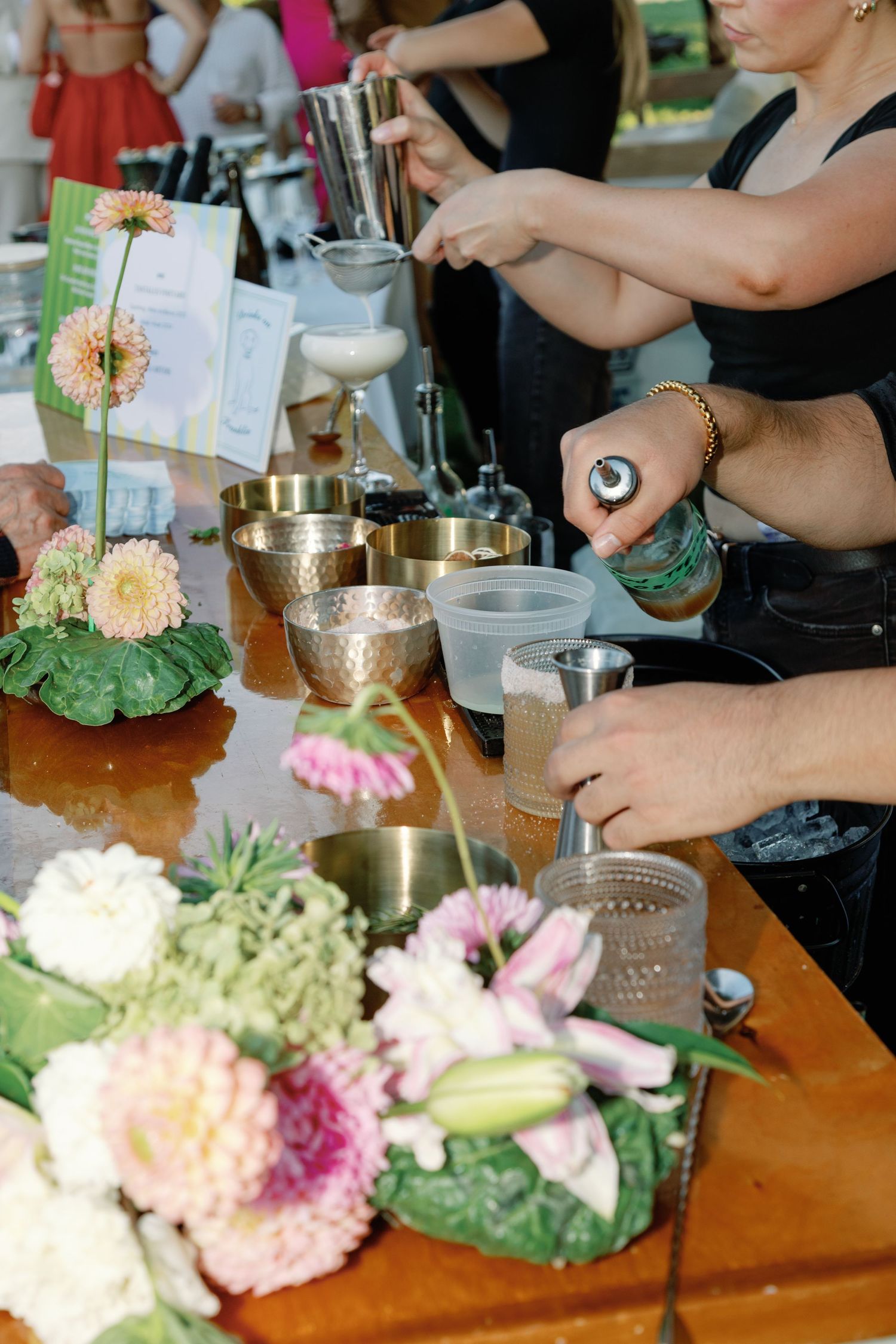 Bartenders prepare cocktails at floral-decorated outdoor wedding bar with gold cups and fresh flowers.