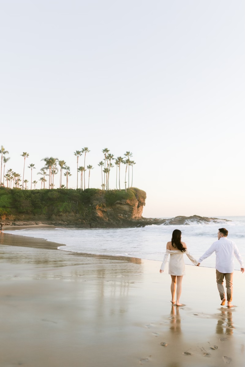 Couple walks barefoot hand in hand along a serene beach at sunset with palm trees behind.