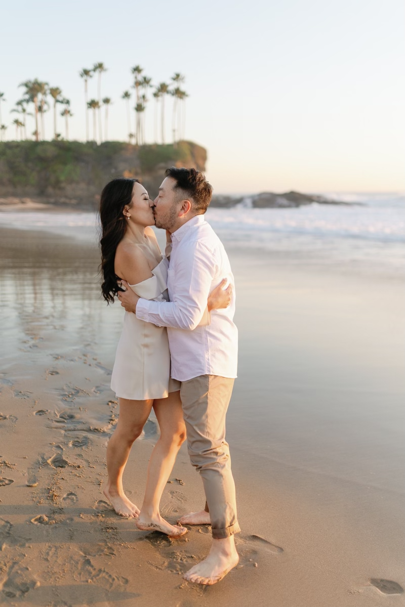 Couple sharing a romantic kiss on a sandy beach at sunset with palm trees in the background.