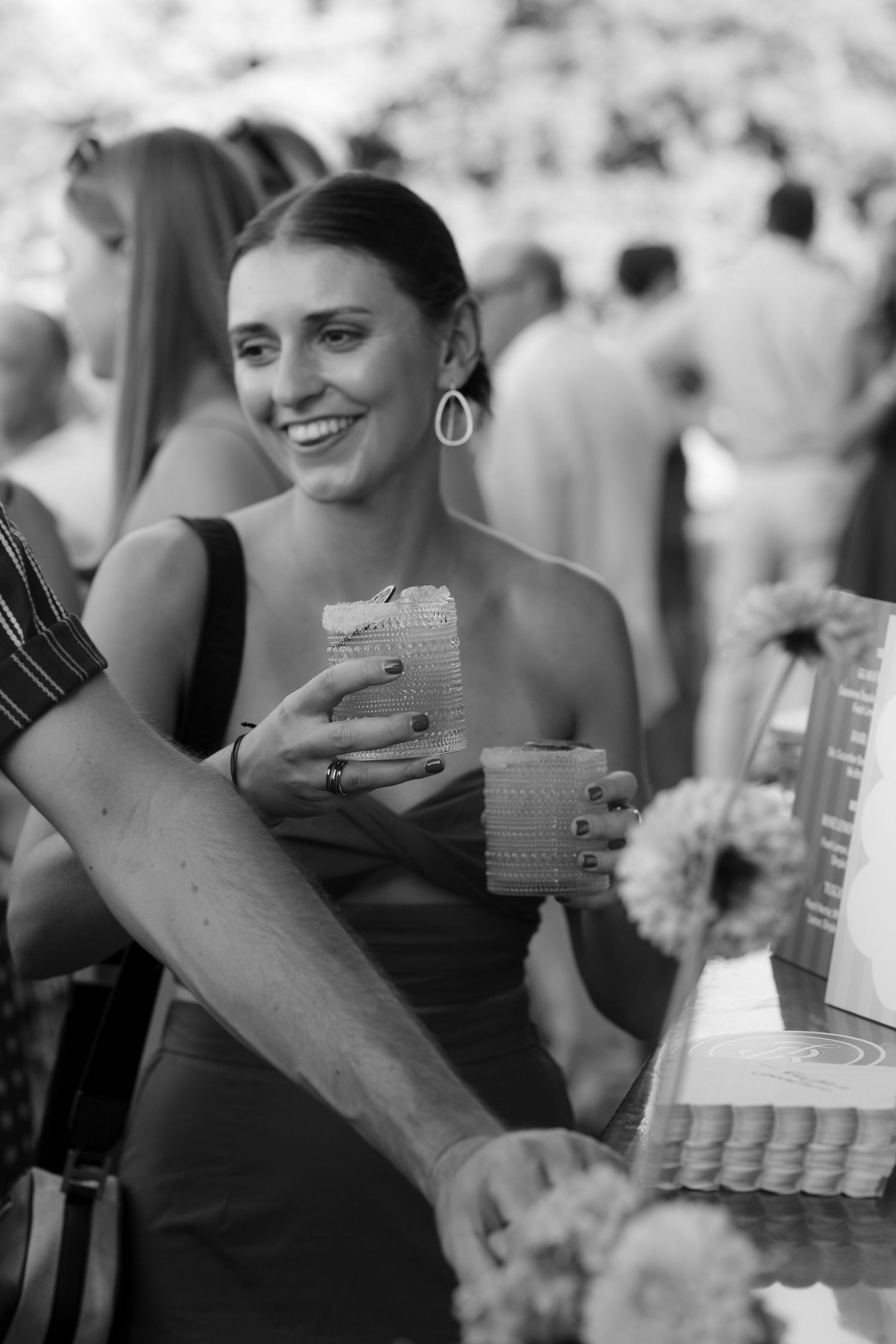 Smiling woman in black dress holds a drink at a lively outdoor wedding reception.