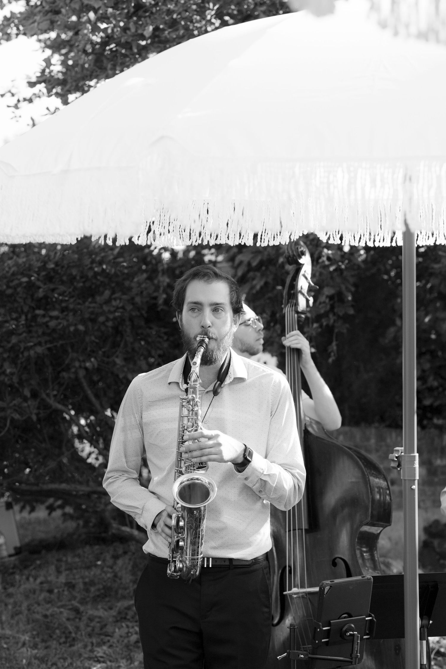 Black and white photo of saxophonist performing at outdoor wedding with double bassist behind.