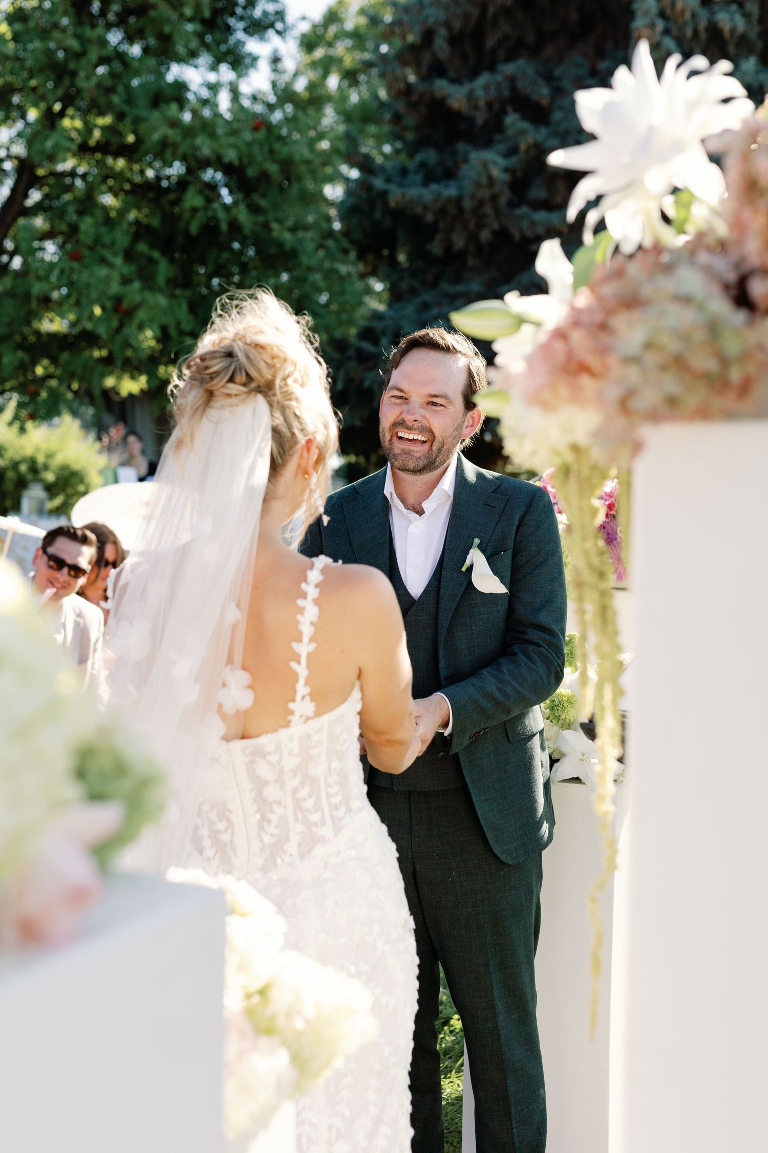 Groom in dark green suit laughing joyfully as bride in lace dress approaches outdoor wedding altar.