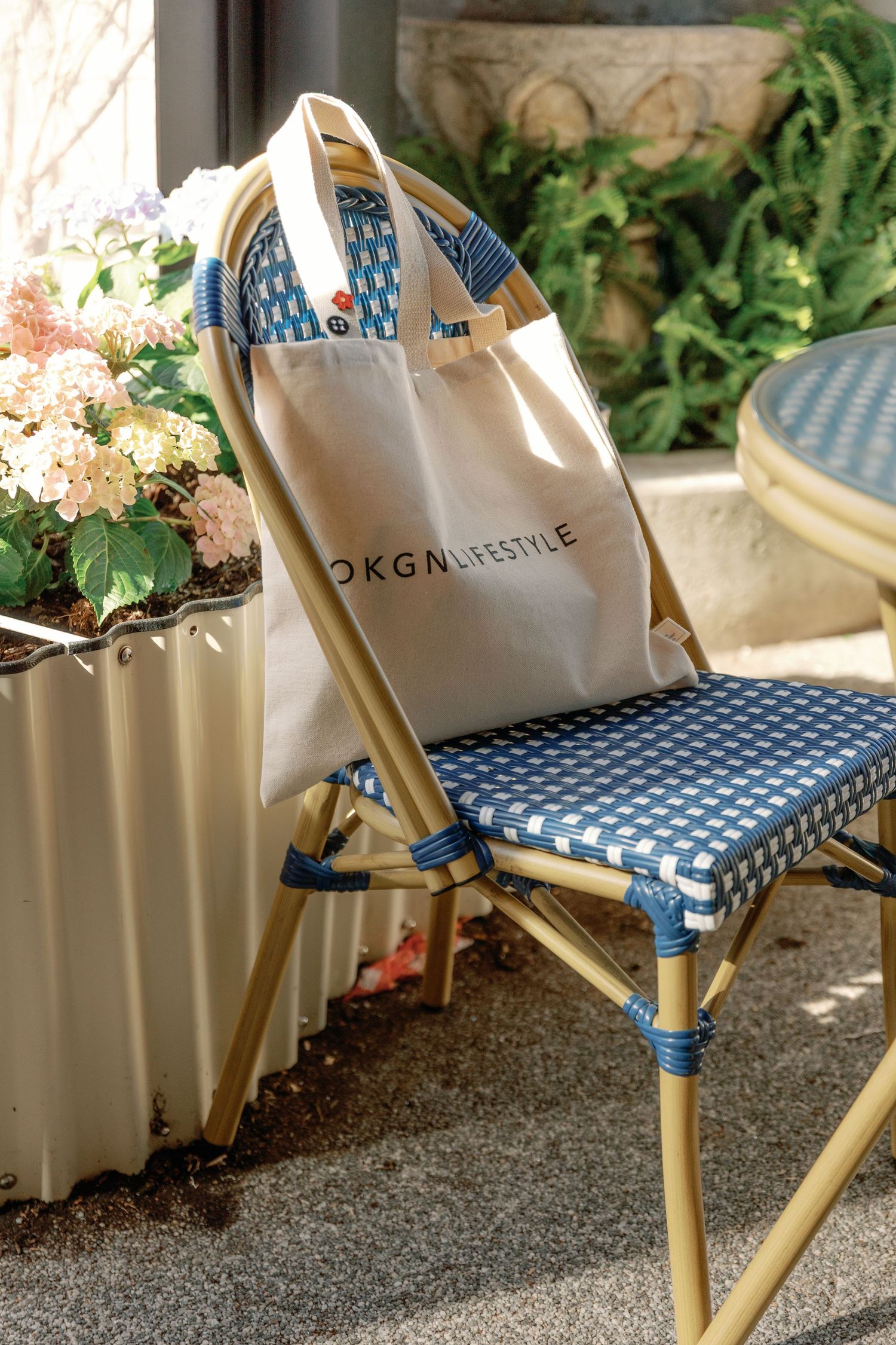 A DKGN Lifestyle tote bag resting on a blue woven bistro chair beside a planter with pink flowers.