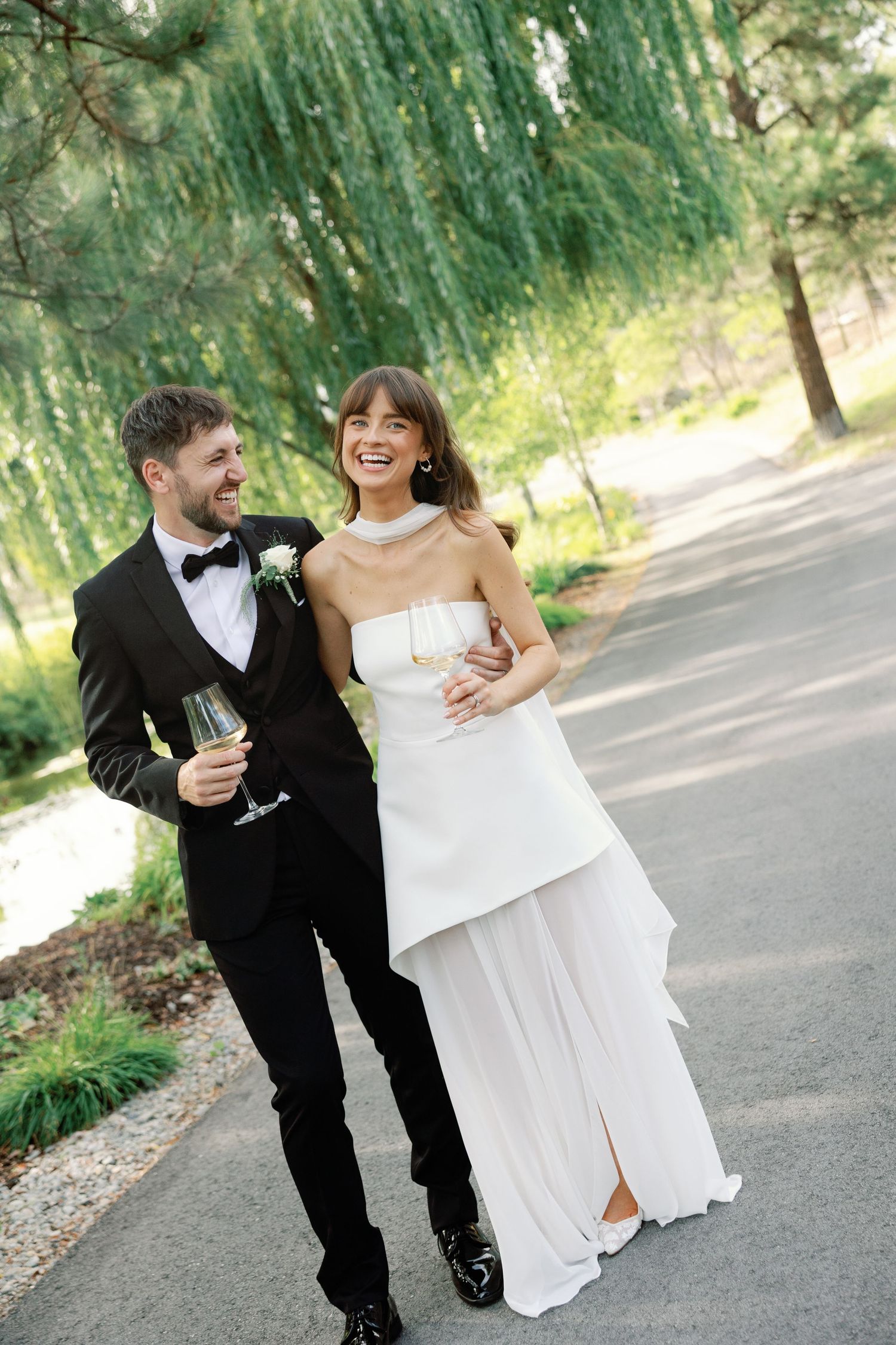 Happy bride and groom walking together in a lush green park, holding champagne glasses on their wedding day.