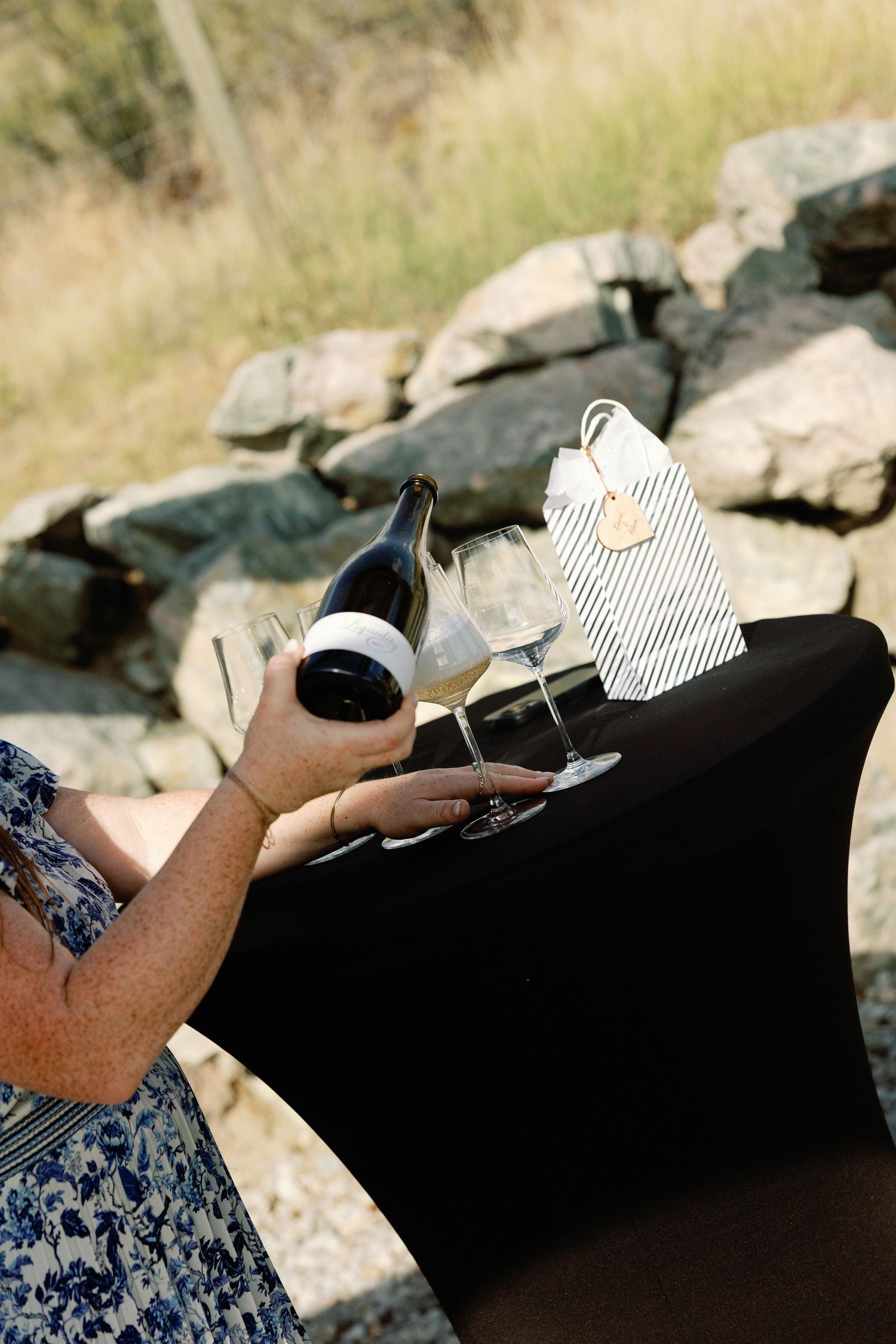 Wedding guest holding wine bottle and glasses at outdoor reception near stone wall.
