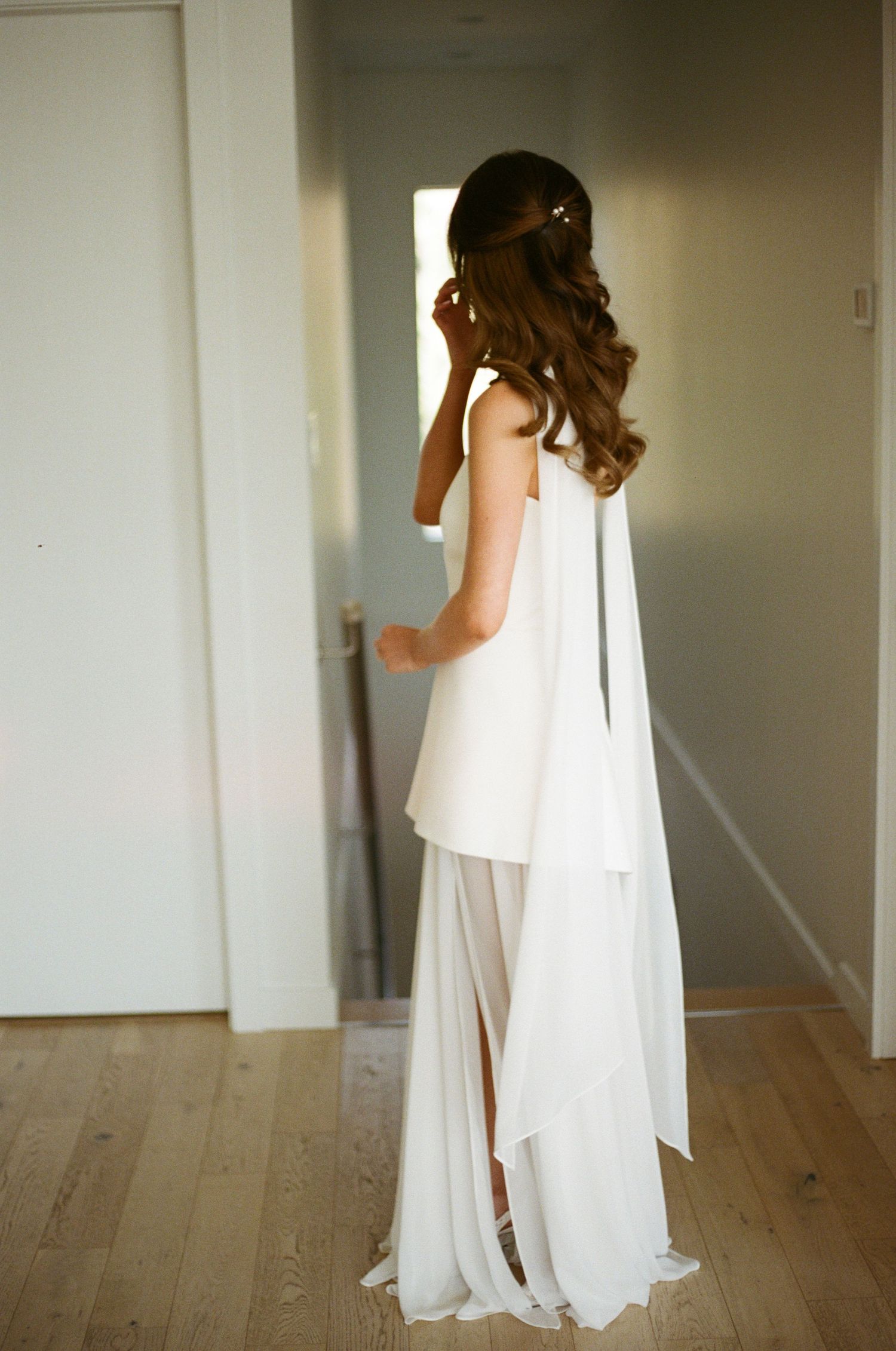 Bride in modern white halter gown with flowing chiffon skirt adjusts hair before mirror.