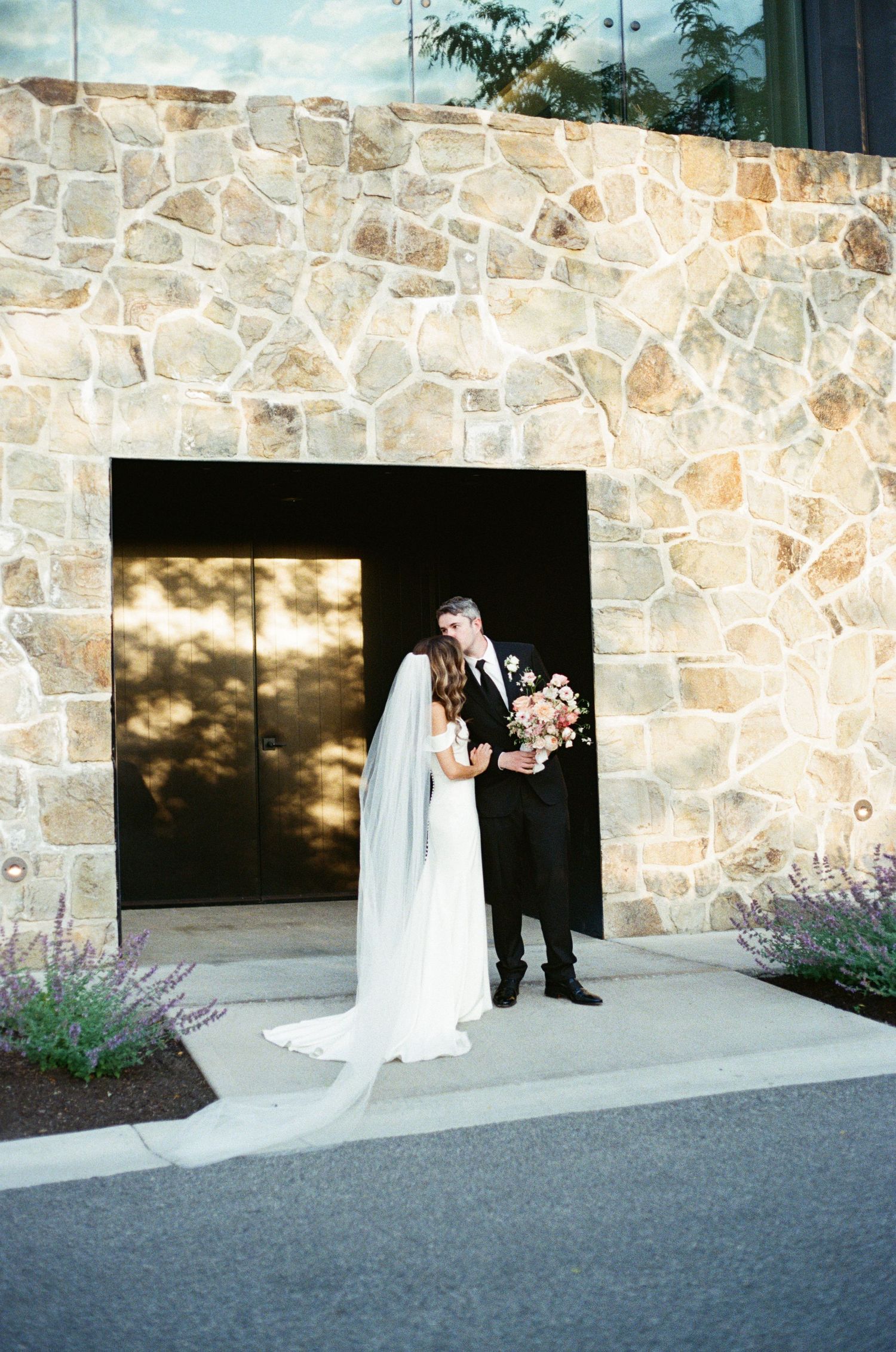 Bride and groom pose before a stone wall entrance, her cathedral veil trailing as he holds a bouquet.
