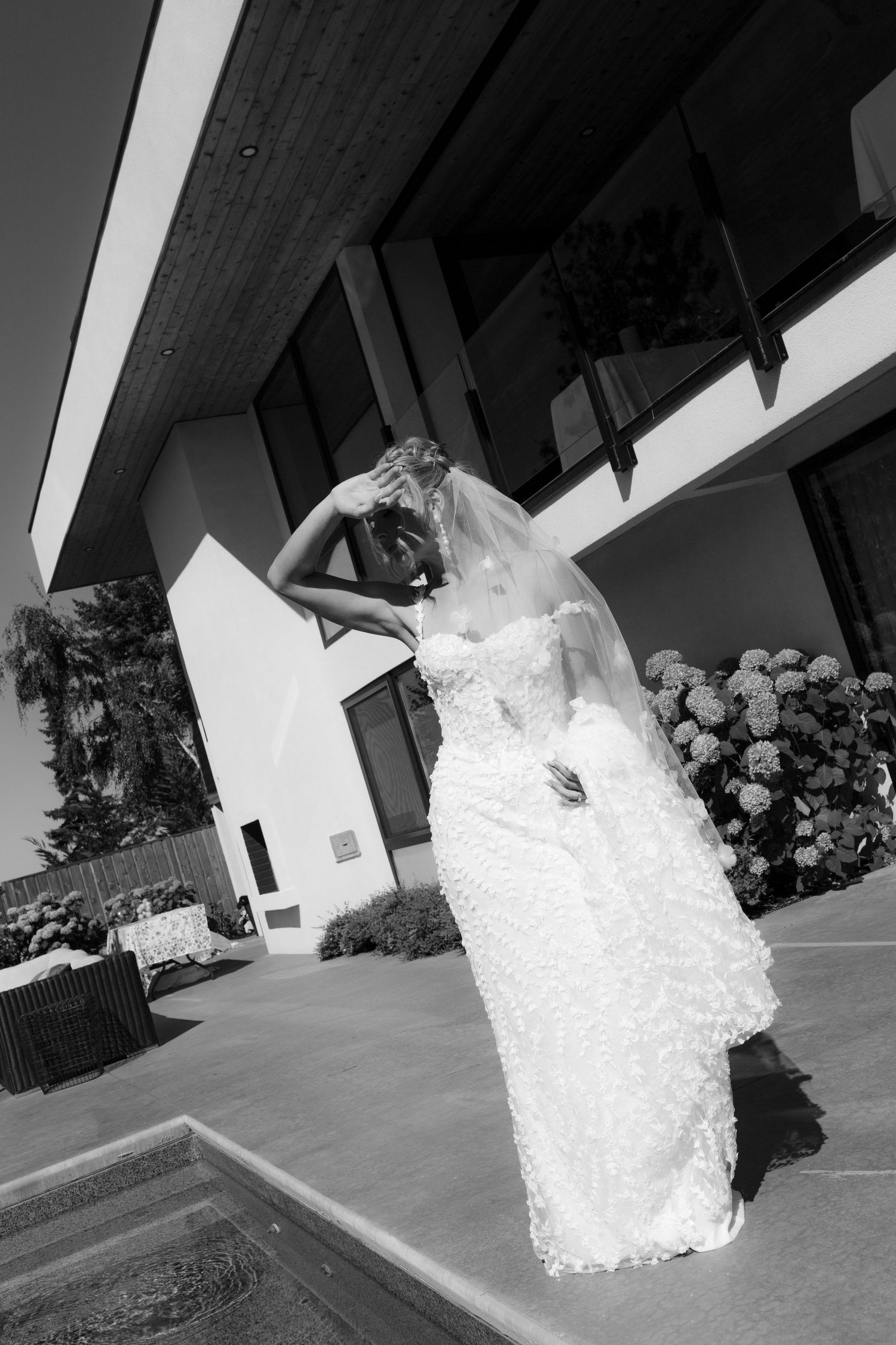 Black and white portrait of bride in lace wedding gown and veil posing outside modern architectural venue.