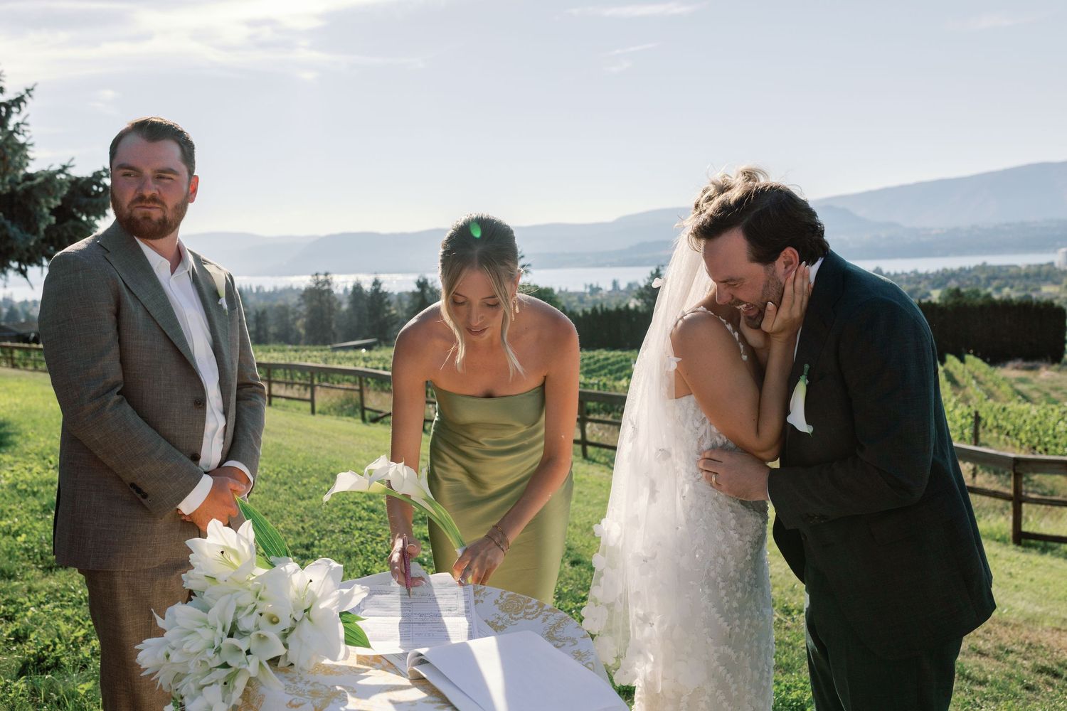 Wedding party signs marriage certificate outdoors with scenic mountain and lake views behind them.