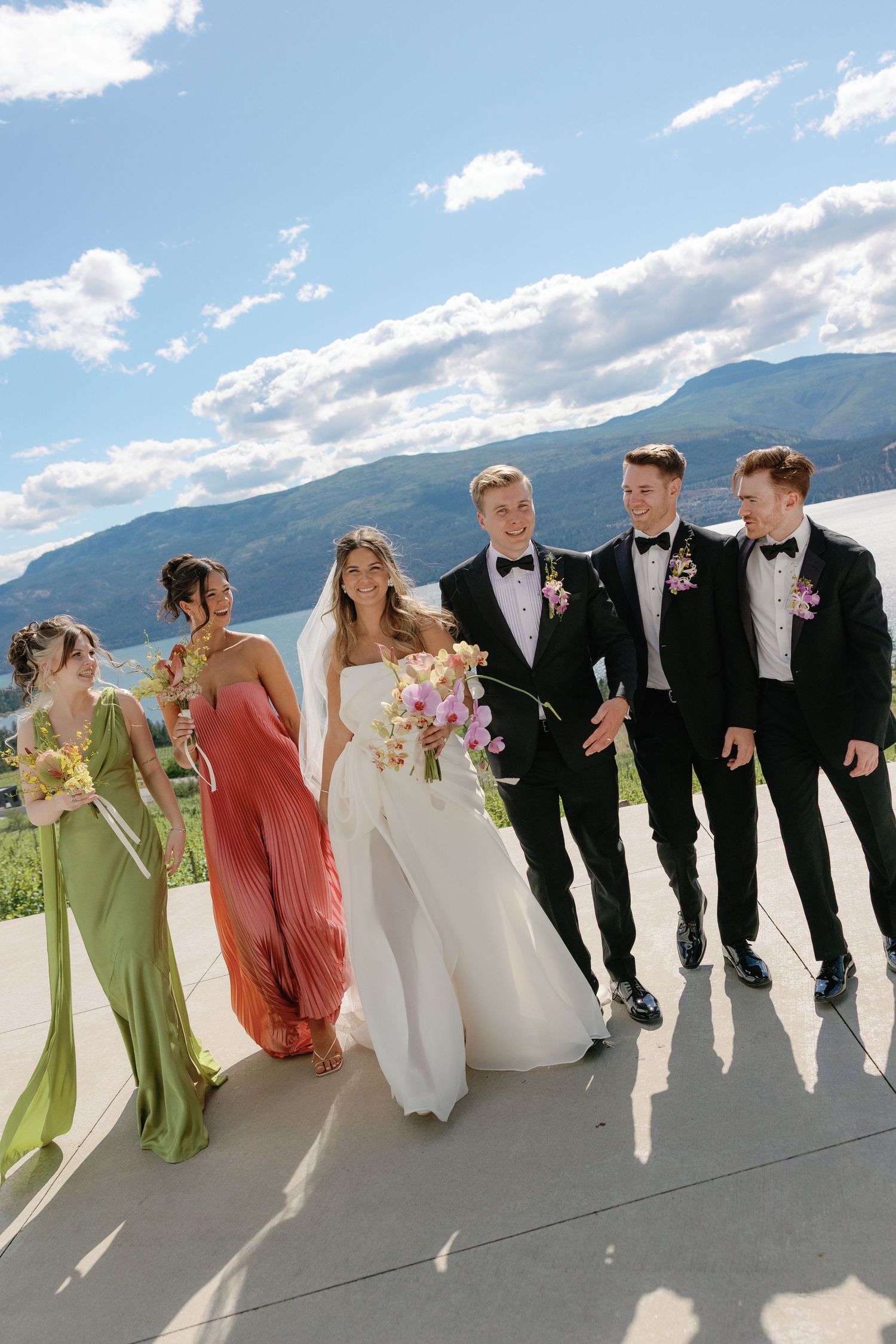 Wedding party in colorful gowns and tuxedos pose outdoors with mountain lake backdrop on sunny day.