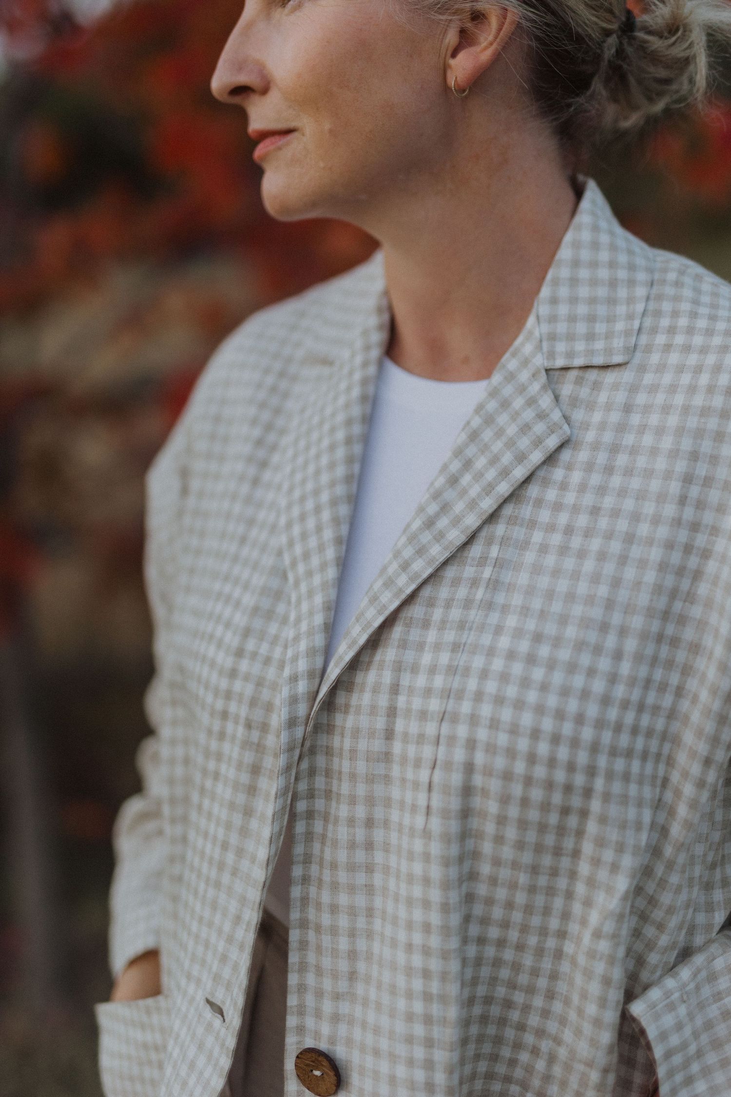 Woman in a beige gingham blazer over white tee, posed outdoors with autumn red foliage background.