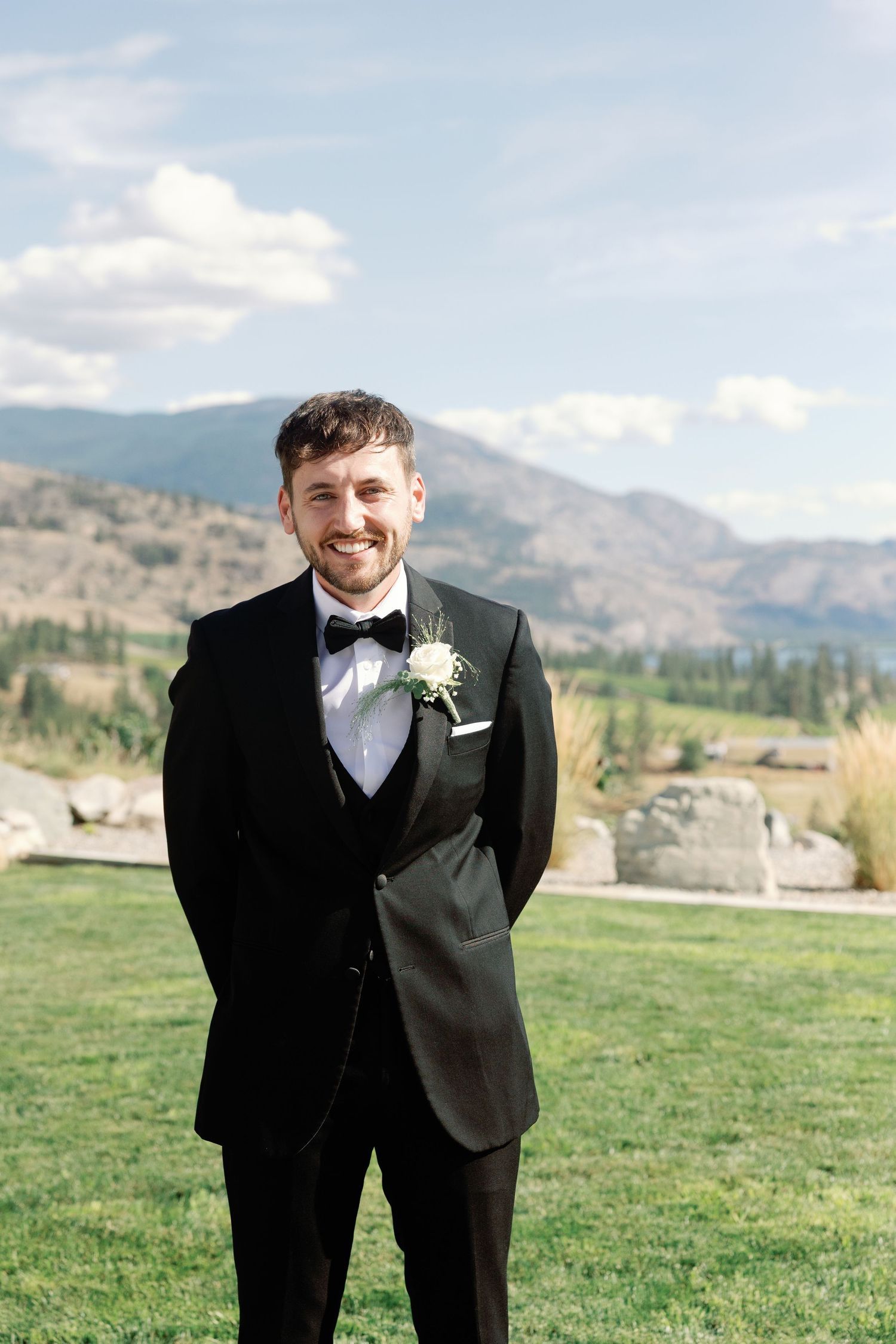 Smiling groom in black tuxedo with boutonniere stands on green lawn with mountain backdrop.